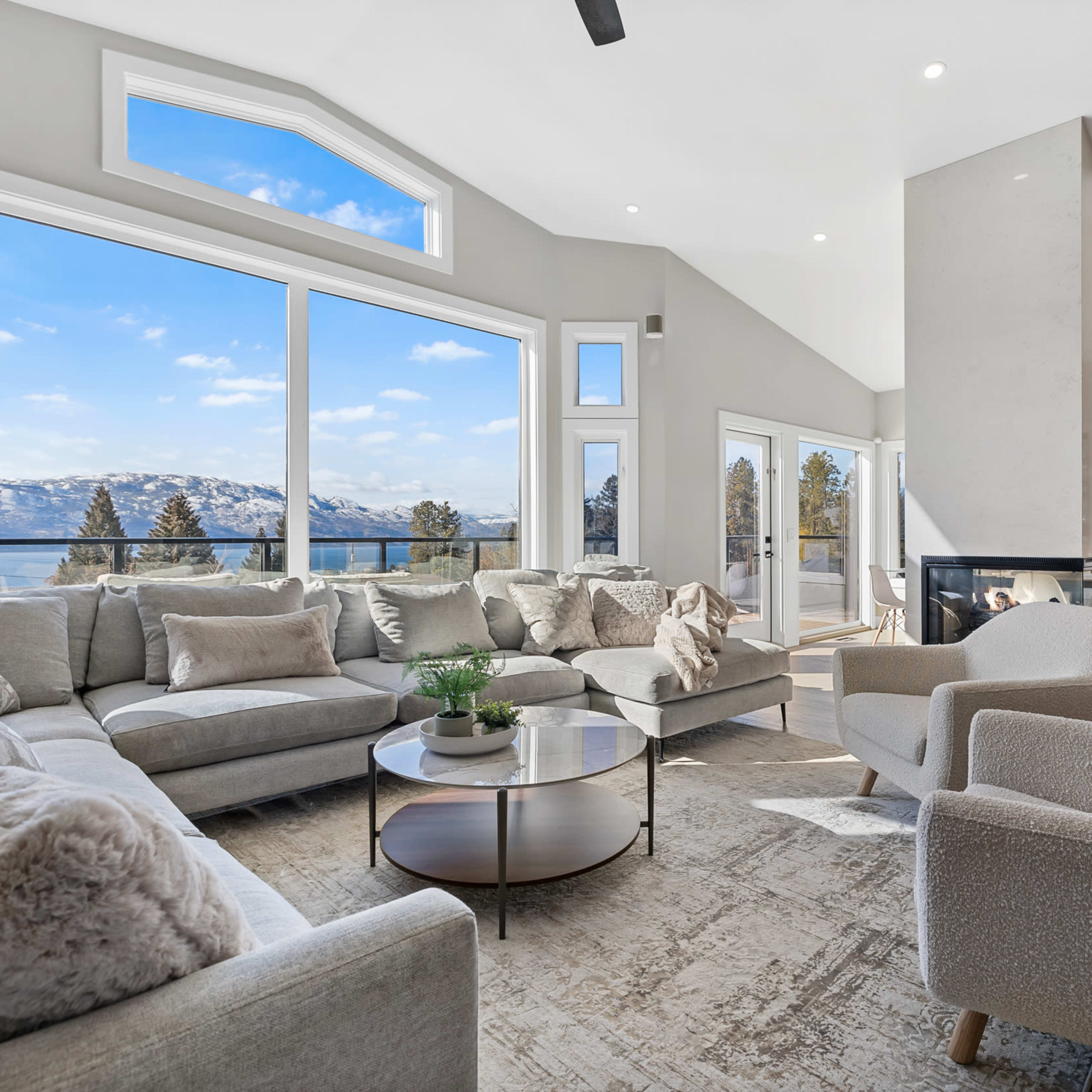 A bright living room with large windows overlooking a lake and mountains, featuring a light gray sectional sofa, a round coffee table, and modern decor.