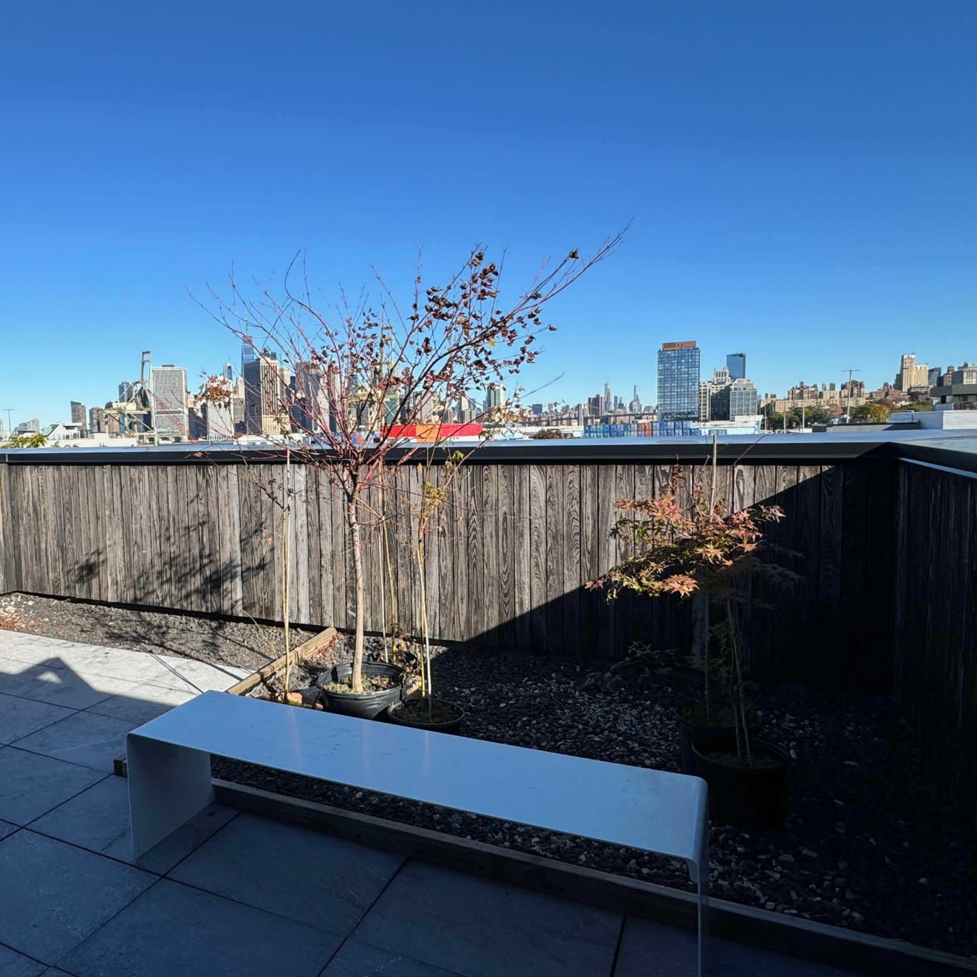 A rooftop terrace features a white bench and small trees, overlooking a city skyline against a clear blue sky.