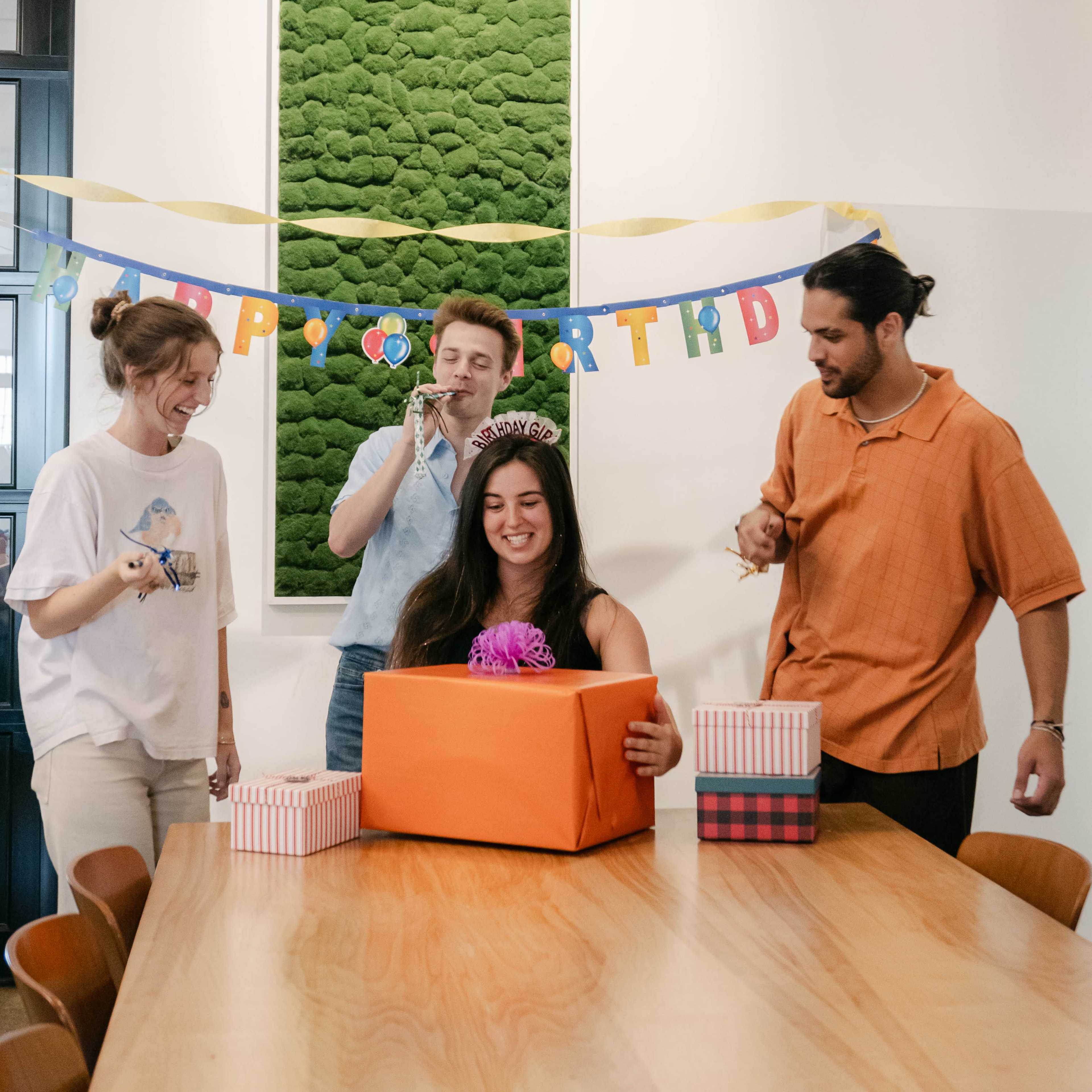 A group of four people gathers around a table in a bright room, celebrating a birthday as one person opens a large orange gift box.