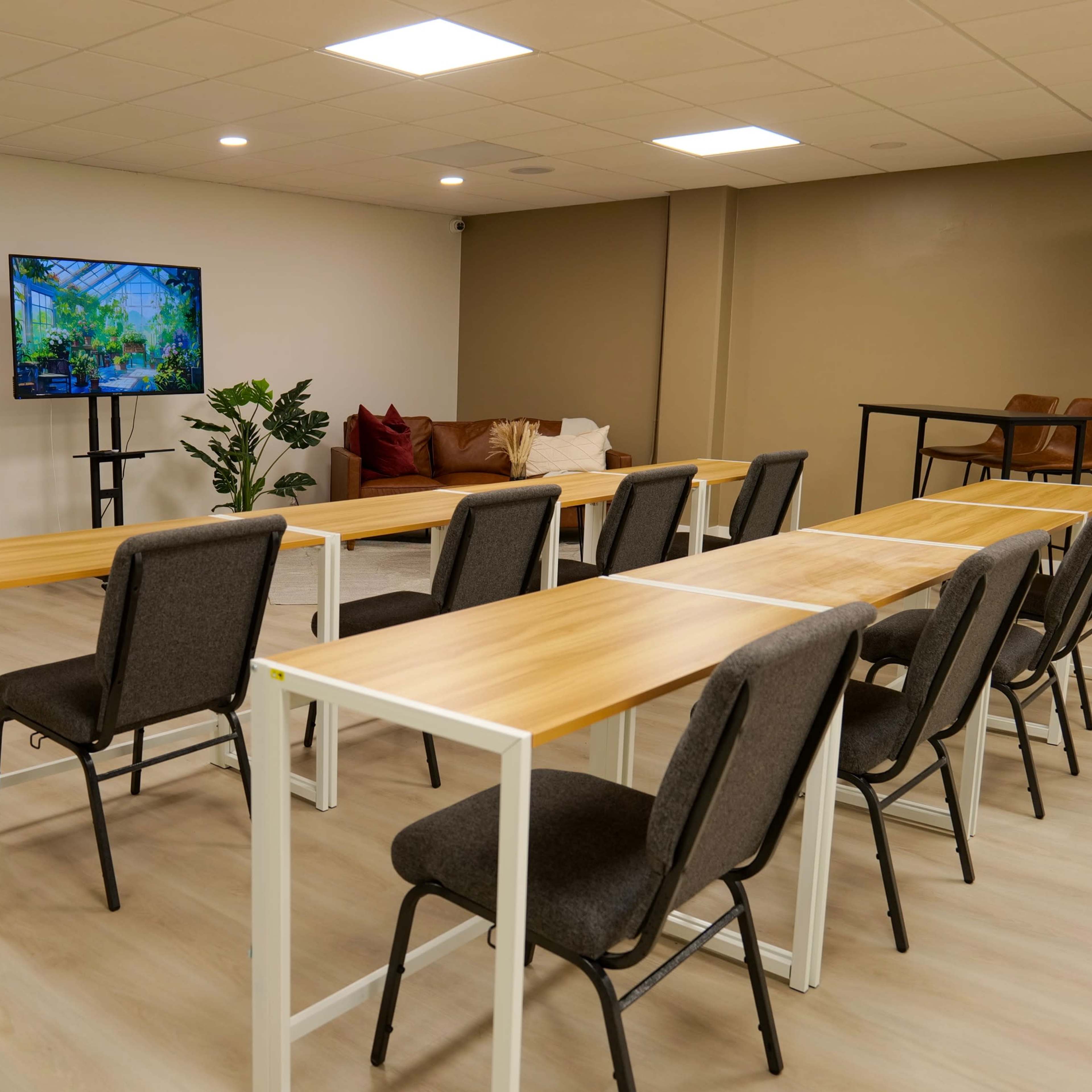 A modern conference room with two screens displaying vibrant images, several long tables arranged in rows, and a seating area with a couch and chairs.