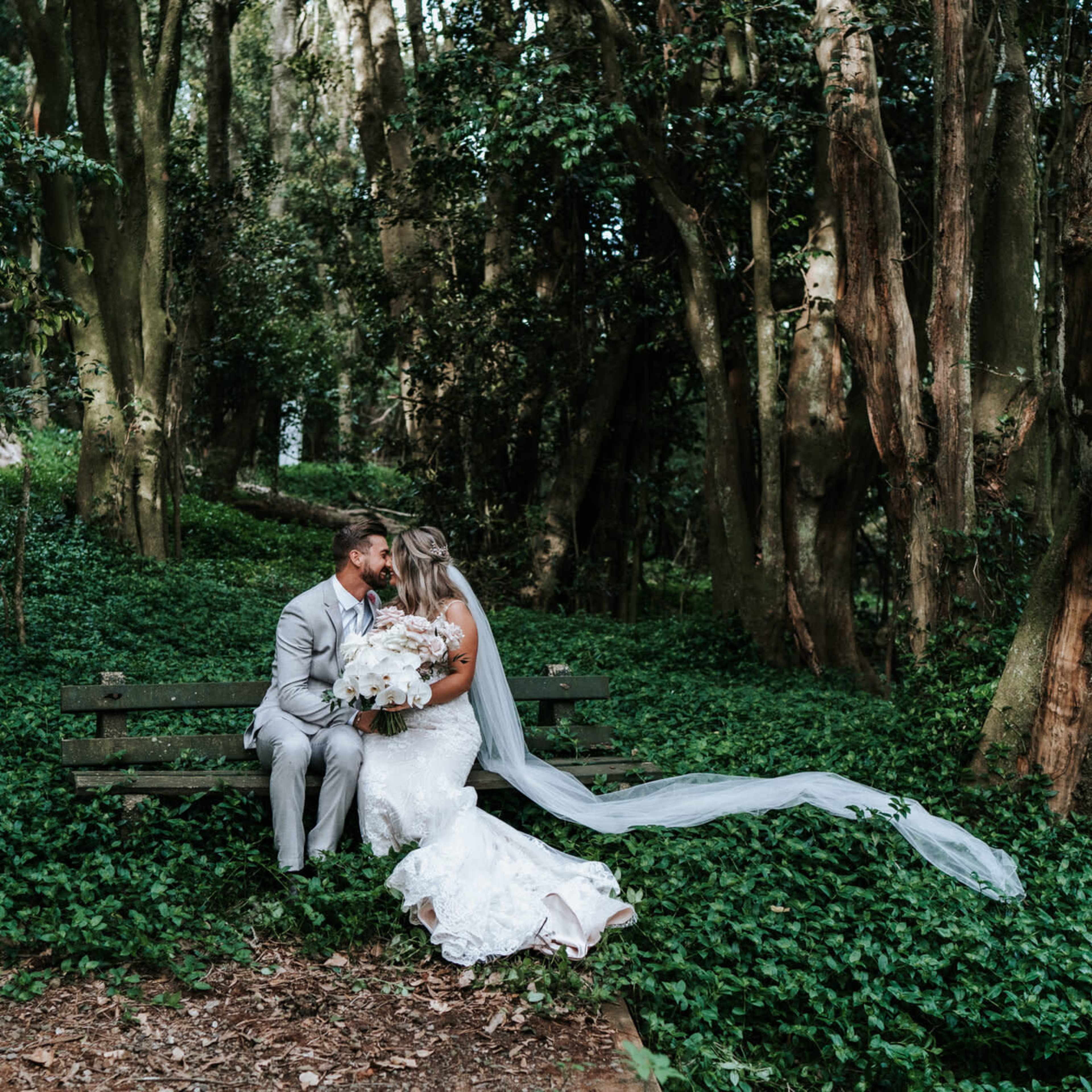A couple sits on a wooden bench in a lush, green forest, surrounded by tall trees and ivy.