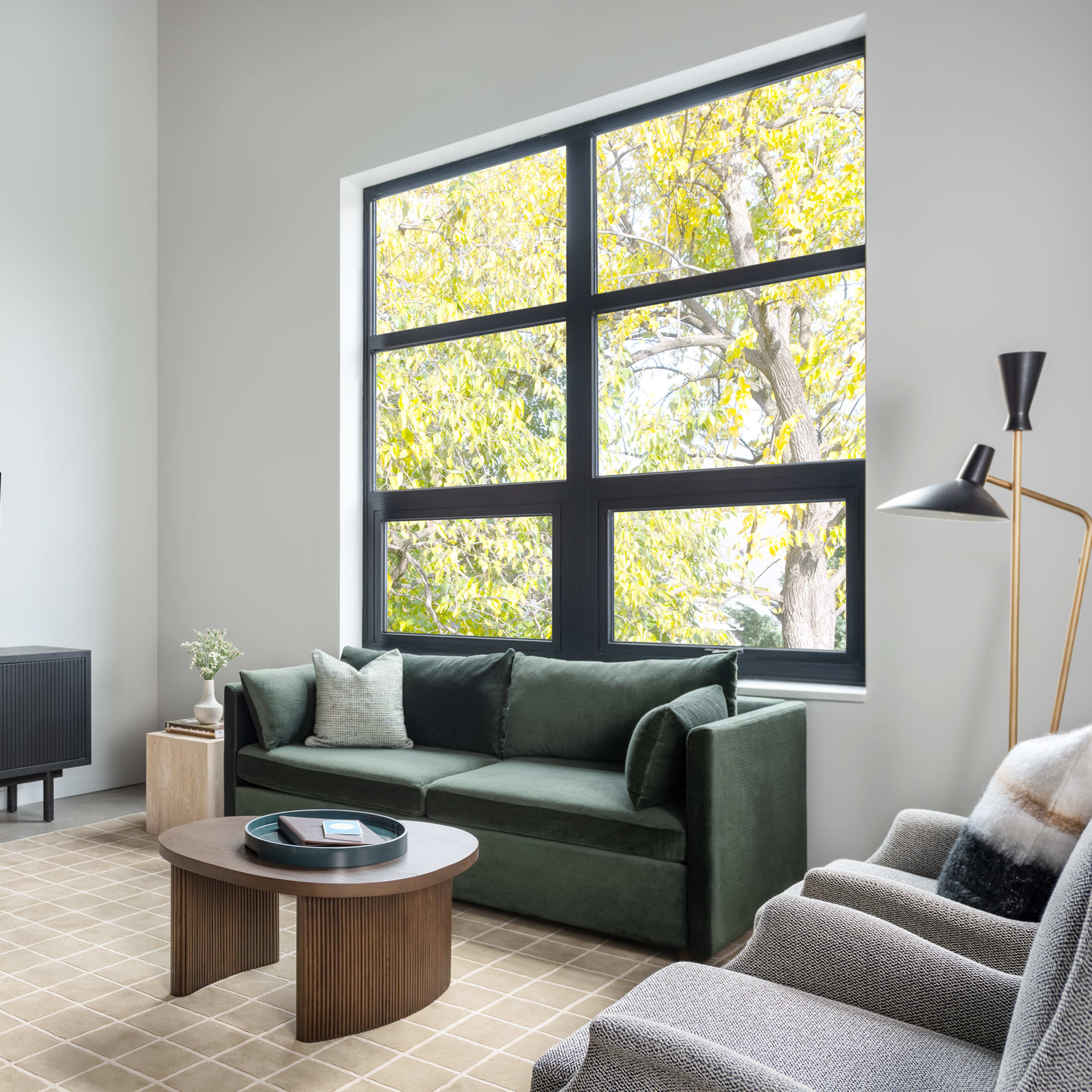 A modern living room features a green sofa, a round table with wooden chairs, and large windows with views of trees, all arranged around a coffee table on a carpet.