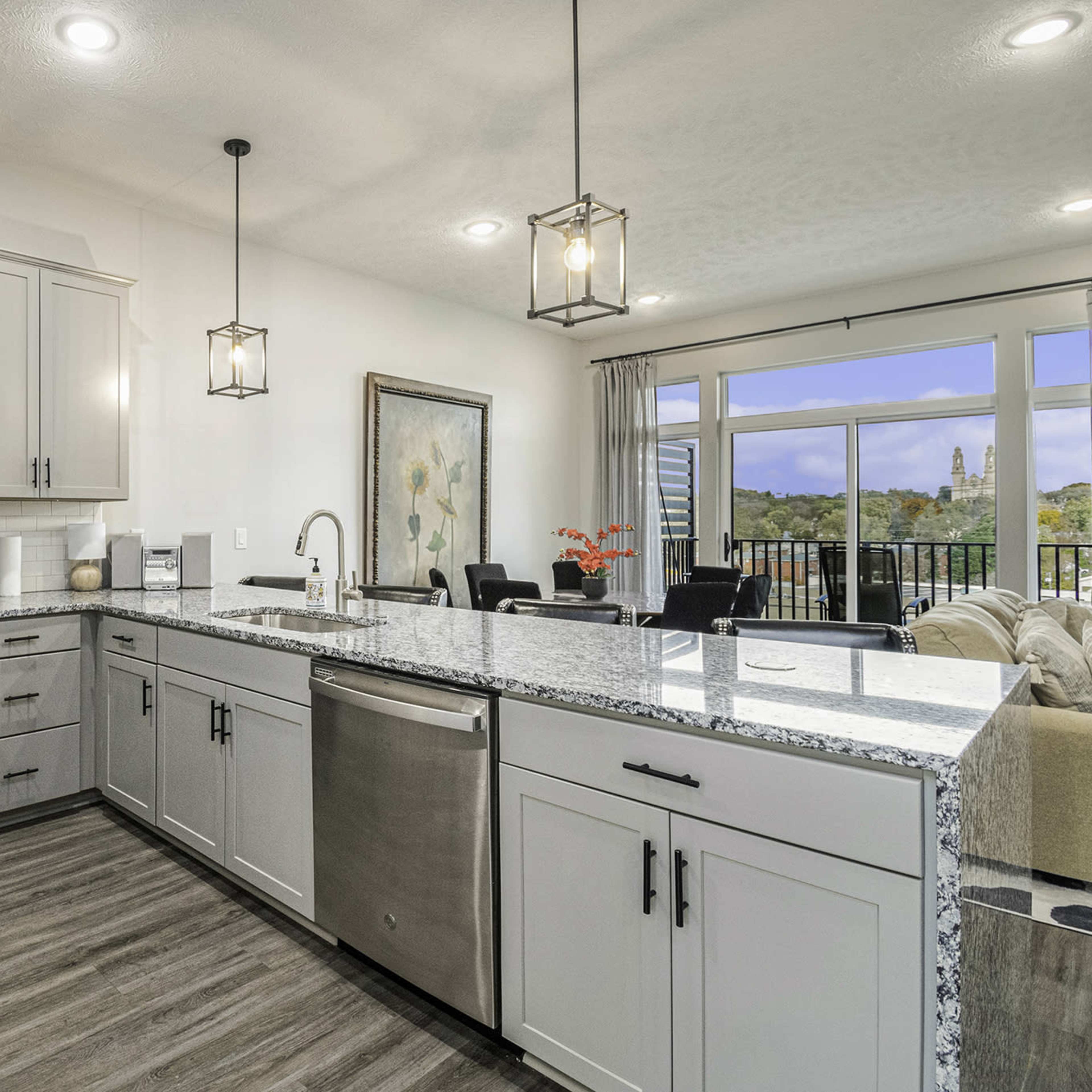 A modern kitchen with stainless steel appliances, gray cabinetry, and a large island with a granite countertop, overlooking a living area with large windows.