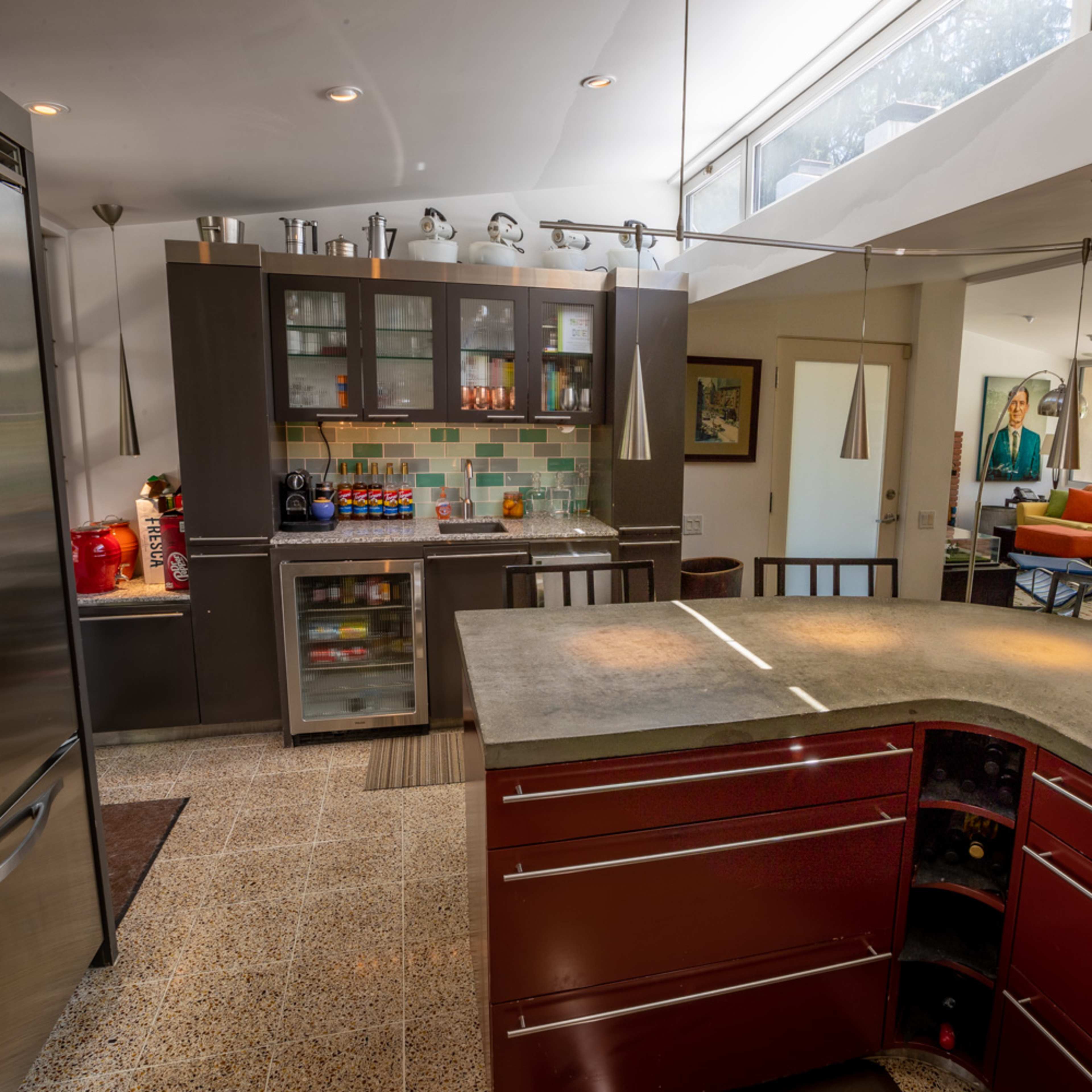 A modern kitchen featuring stainless steel appliances, a concrete countertop island, and a colorful tiled backsplash, with a living area visible in the background.