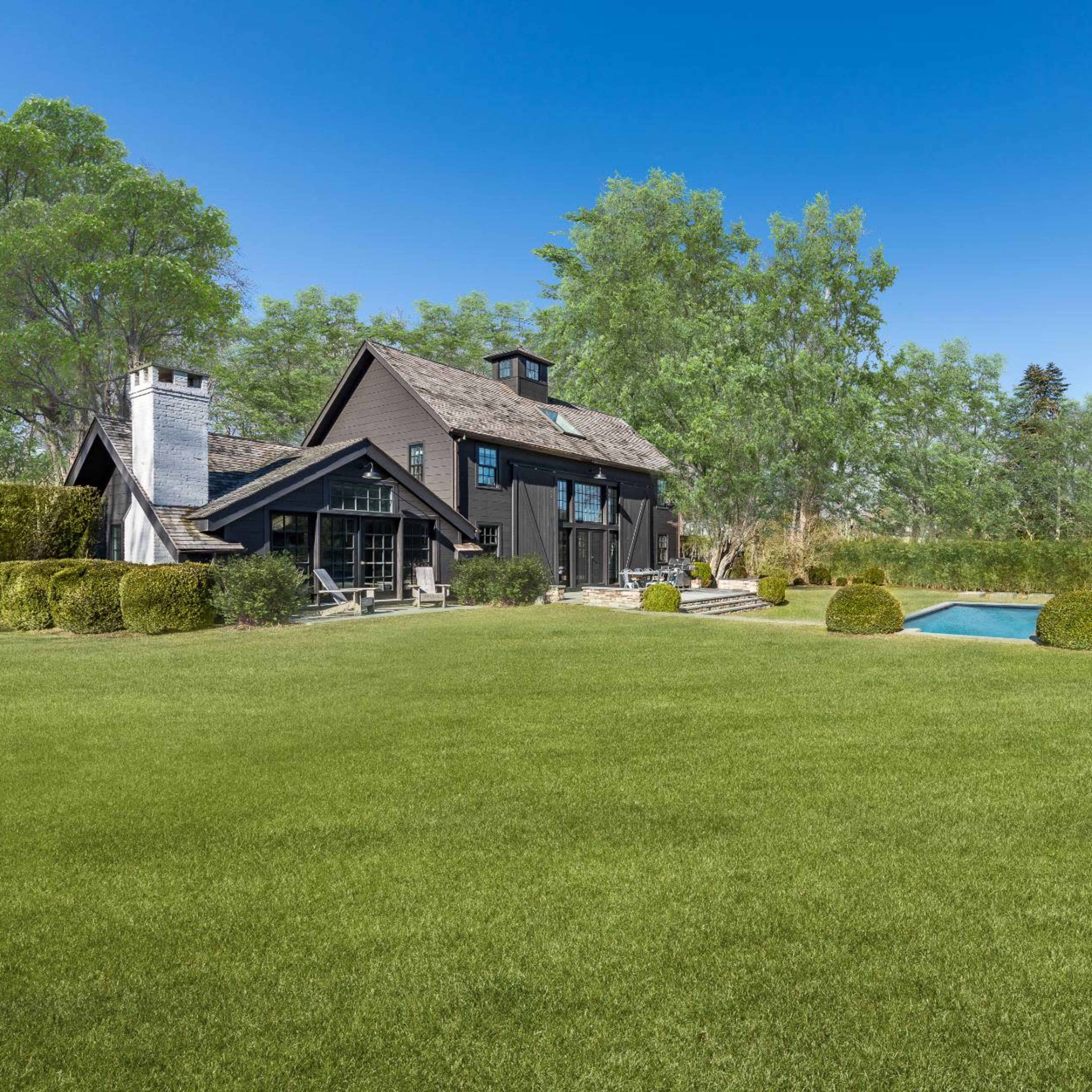 A modern black farmhouse is set in a lush green yard featuring a swimming pool and neatly trimmed hedges.