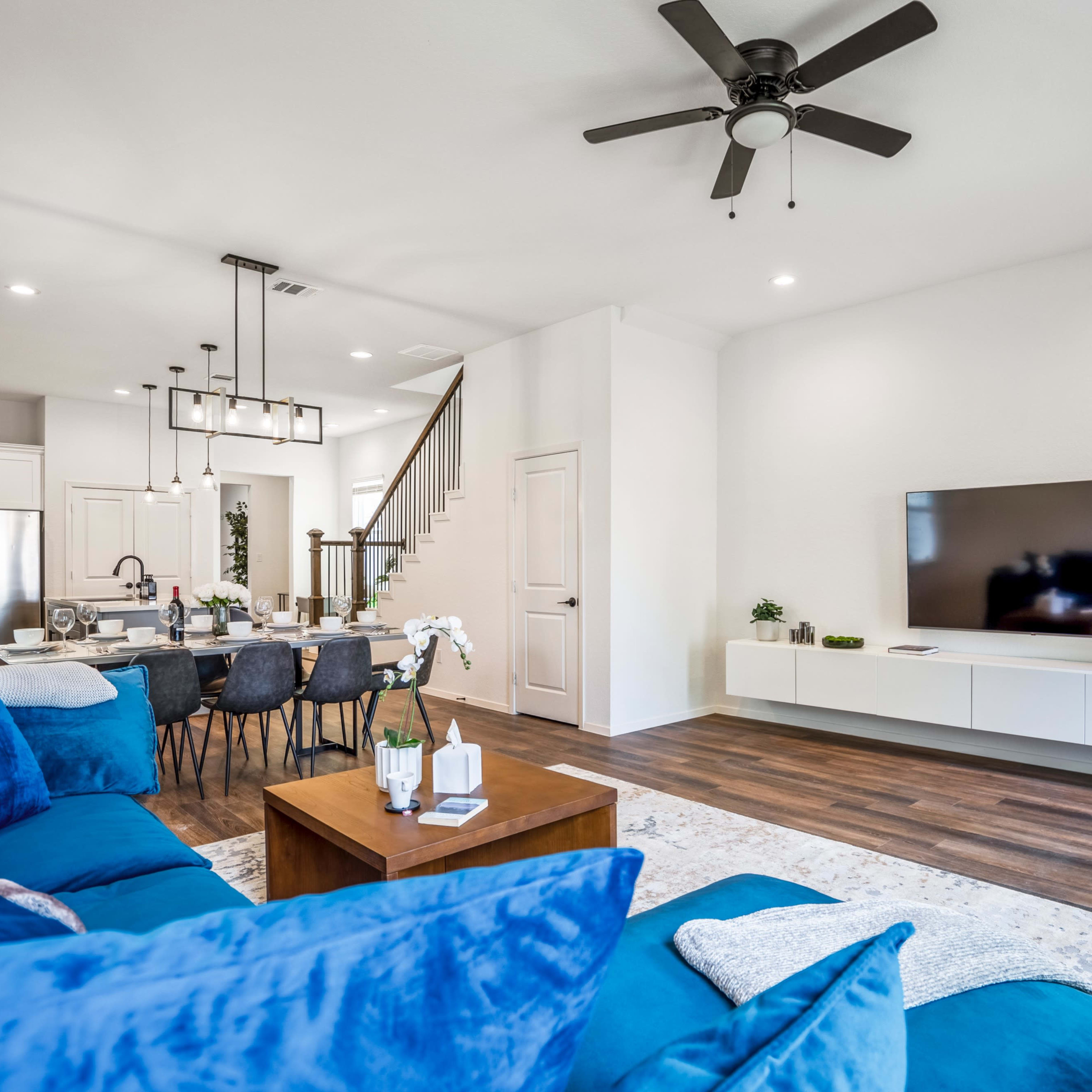 A modern living room features a blue sectional sofa, a wooden coffee table, and a dining area with a large table and chairs, connected to a kitchen with white cabinetry.