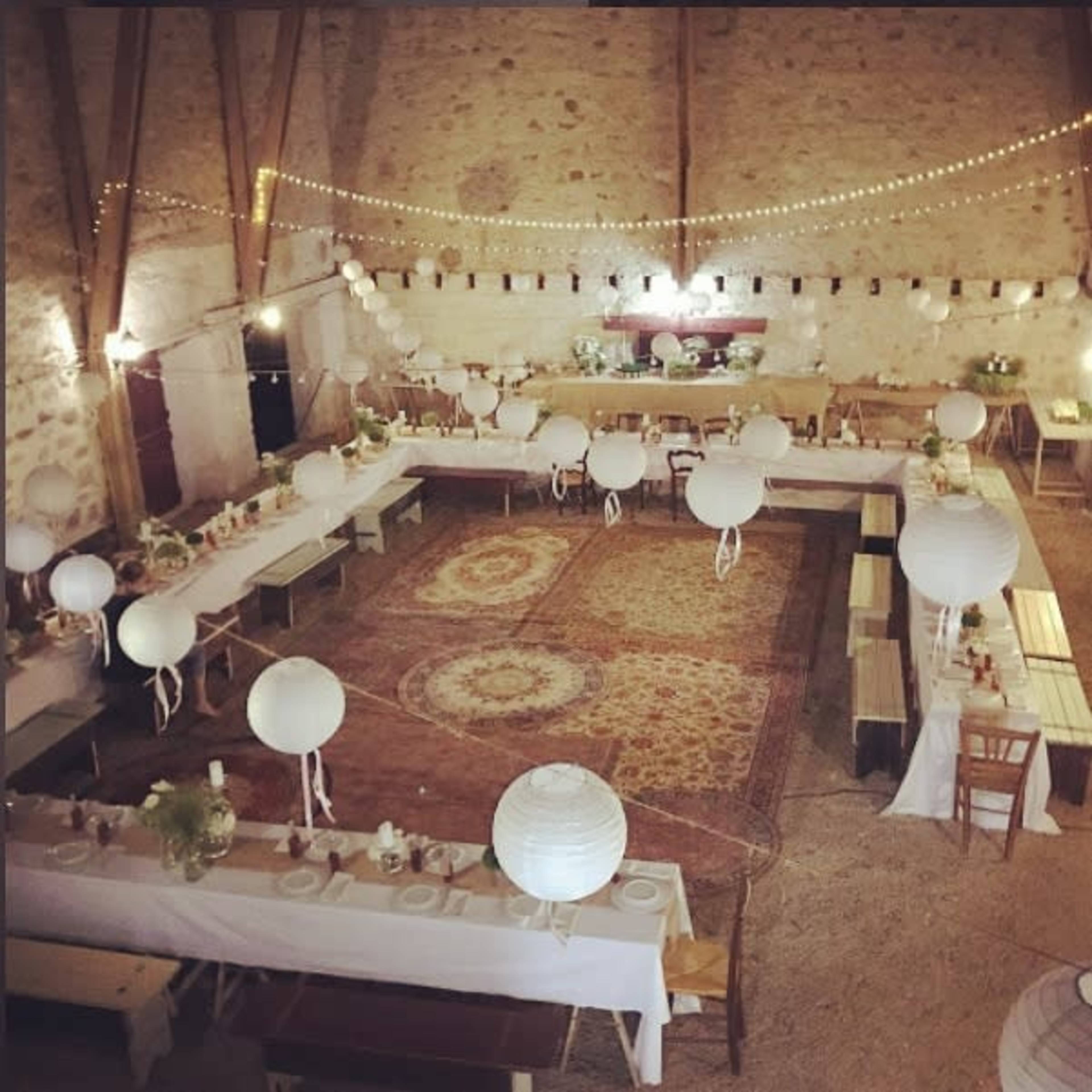 A spacious indoor venue set up for a dining event, featuring long tables adorned with white tablecloths and decorated with paper lanterns and greenery.