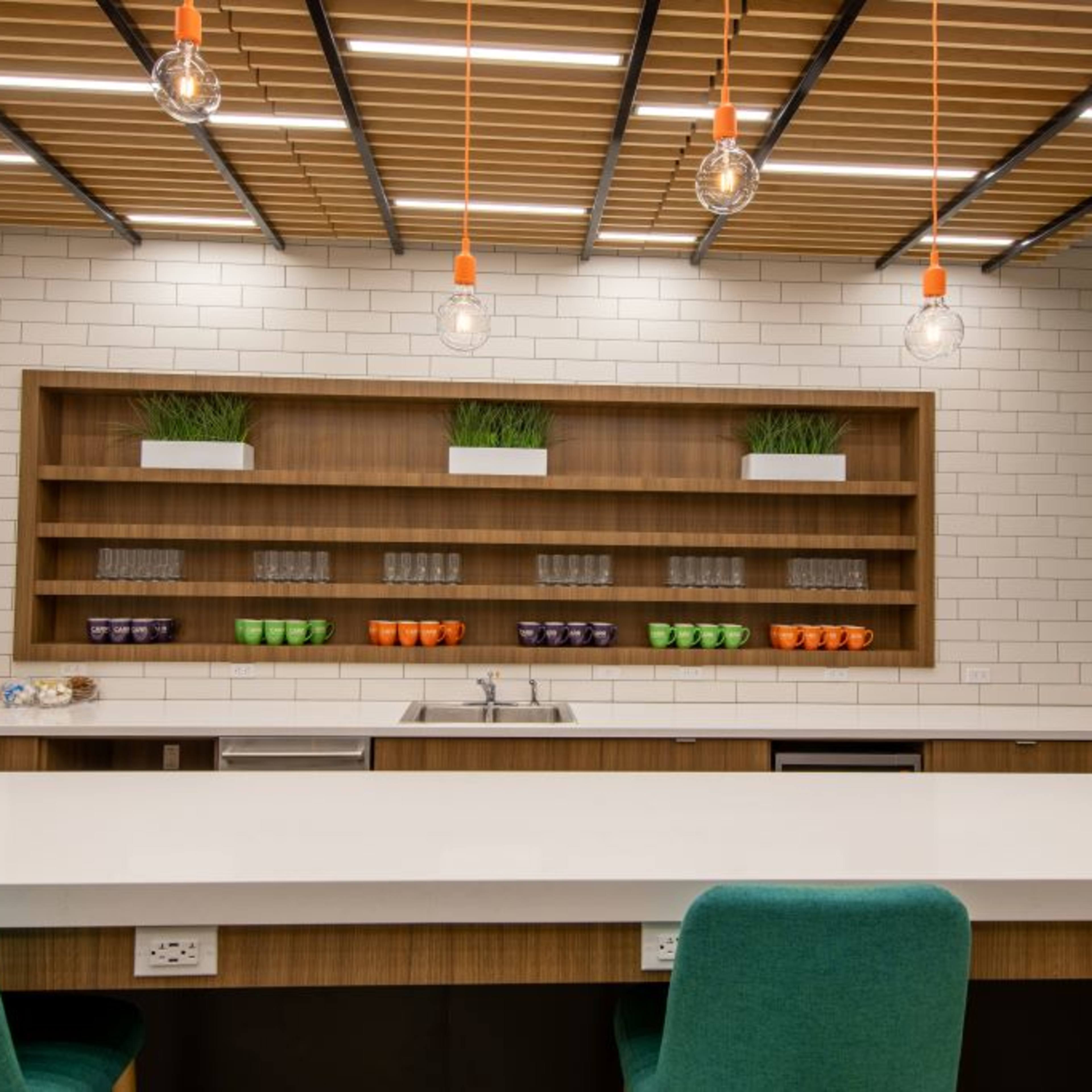 The modern kitchen features a long white countertop, overhead lighting with orange accents, and neatly arranged shelves displaying glasses and colorful cups.