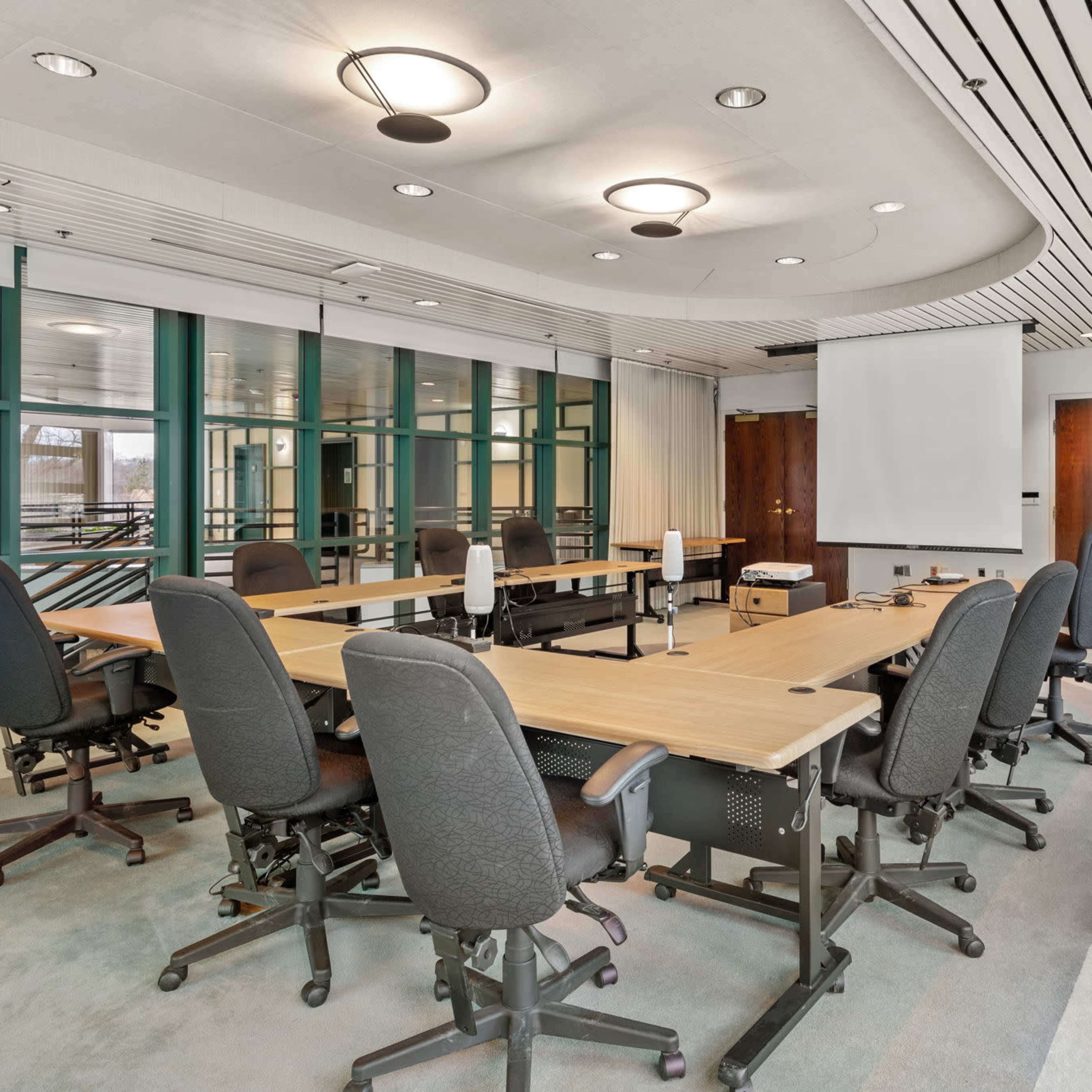 A meeting room features a large, oval table surrounded by ergonomic chairs, with a projector screen at one end and large windows along the walls.