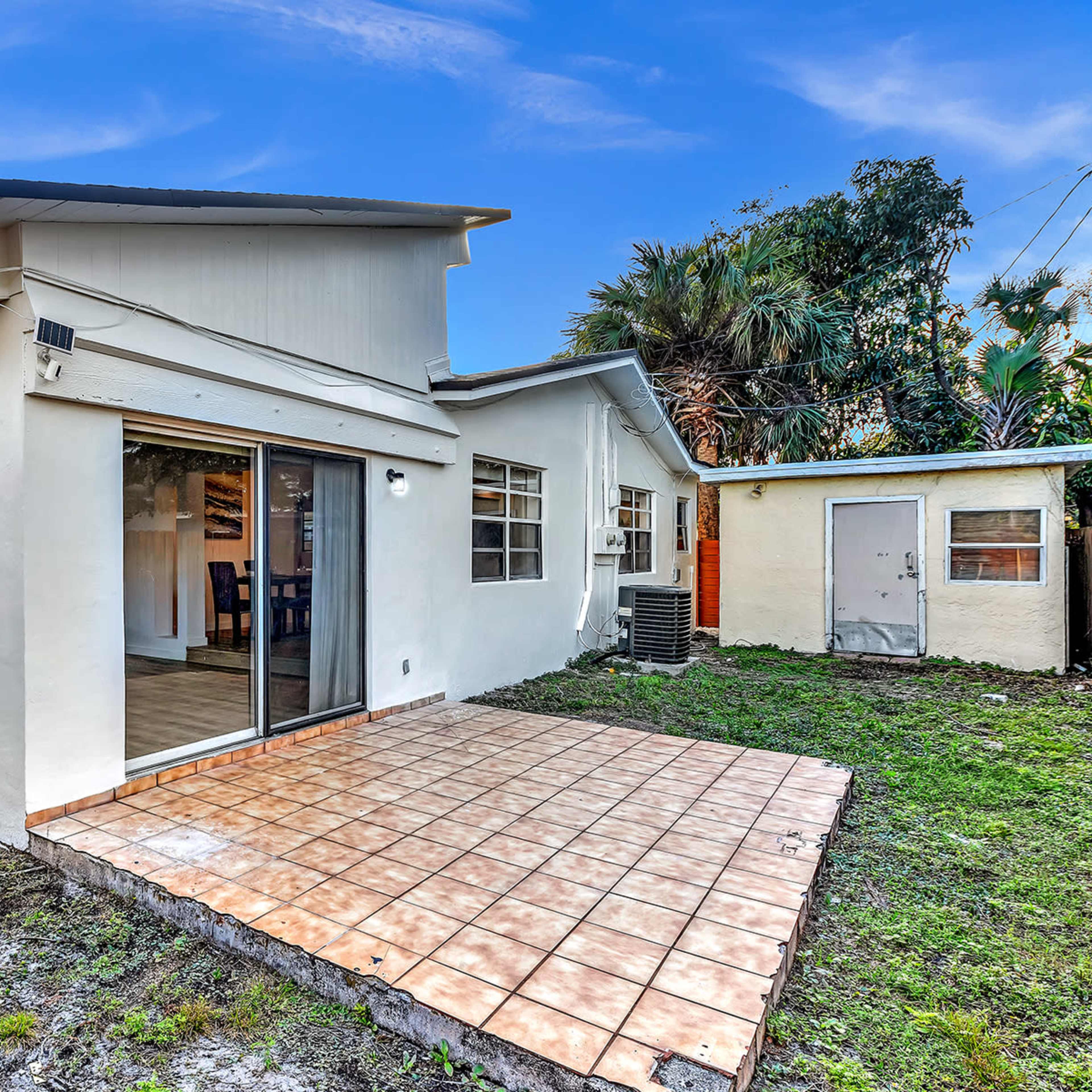 A single-story house with a tiled patio and large sliding glass doors overlooks a grassy yard.