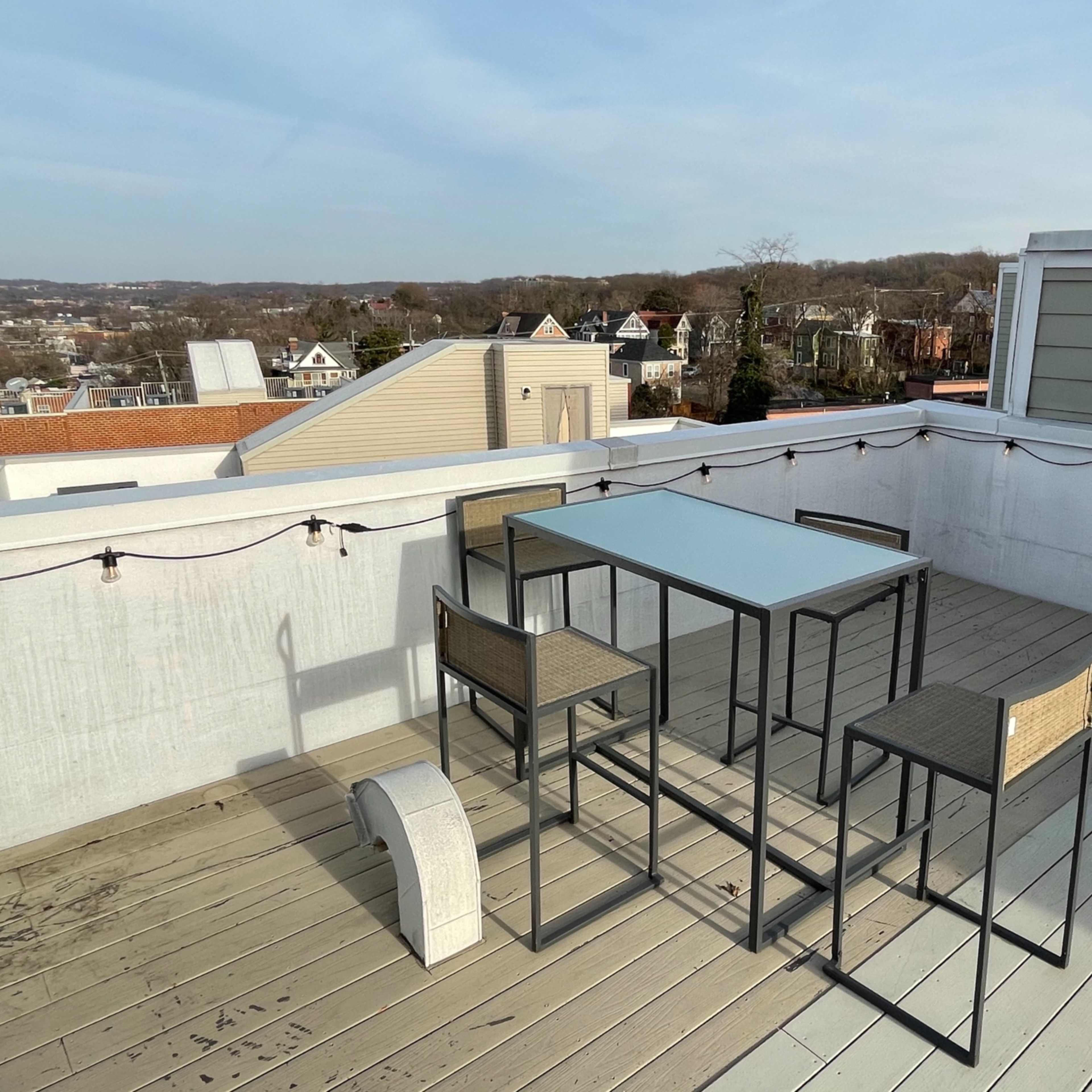 A rooftop patio features a glass-topped table surrounded by four metal chairs, with a view of nearby buildings and a clear sky.