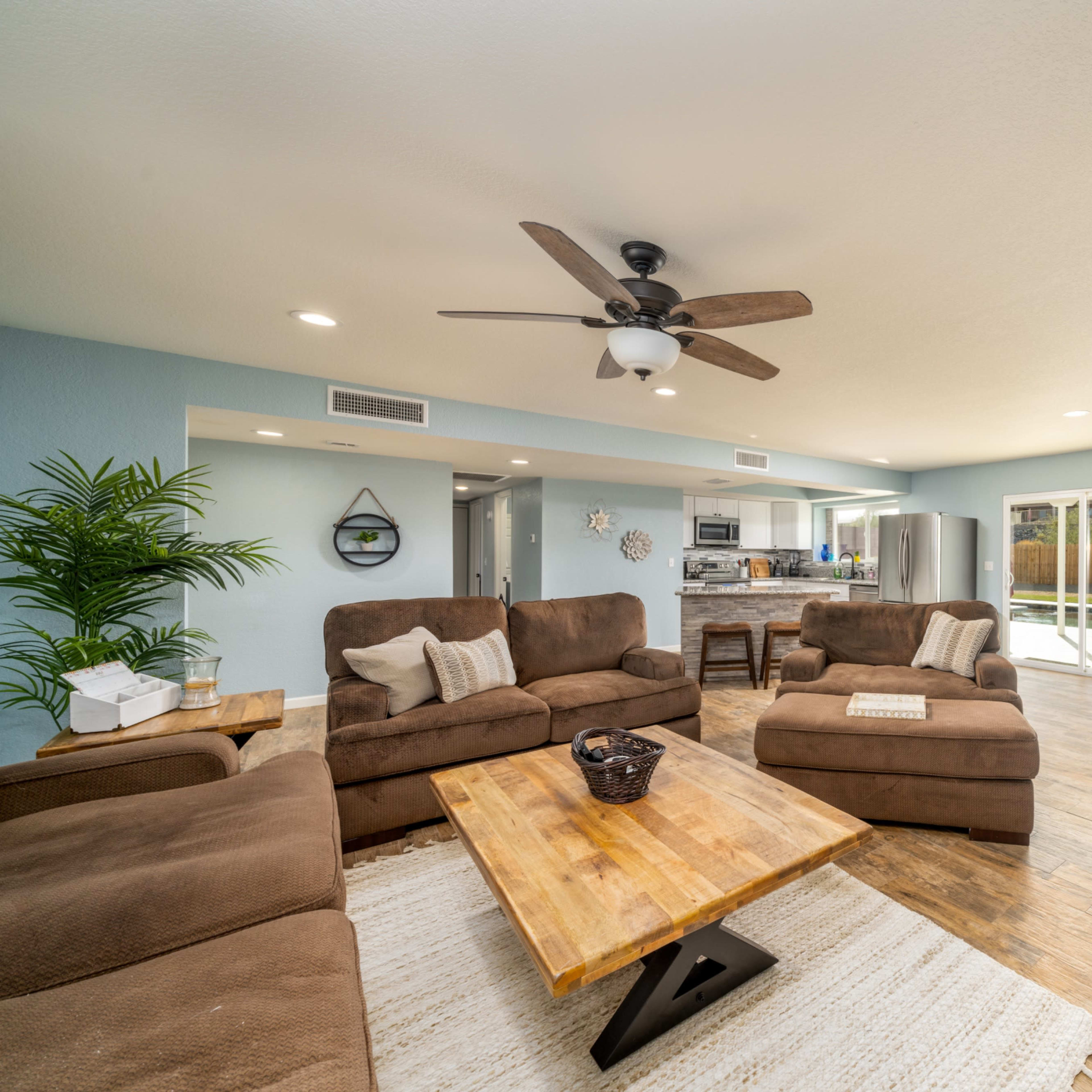 A spacious living room features brown sofas, wooden tables, a dining area, and large windows overlooking an outdoor space.