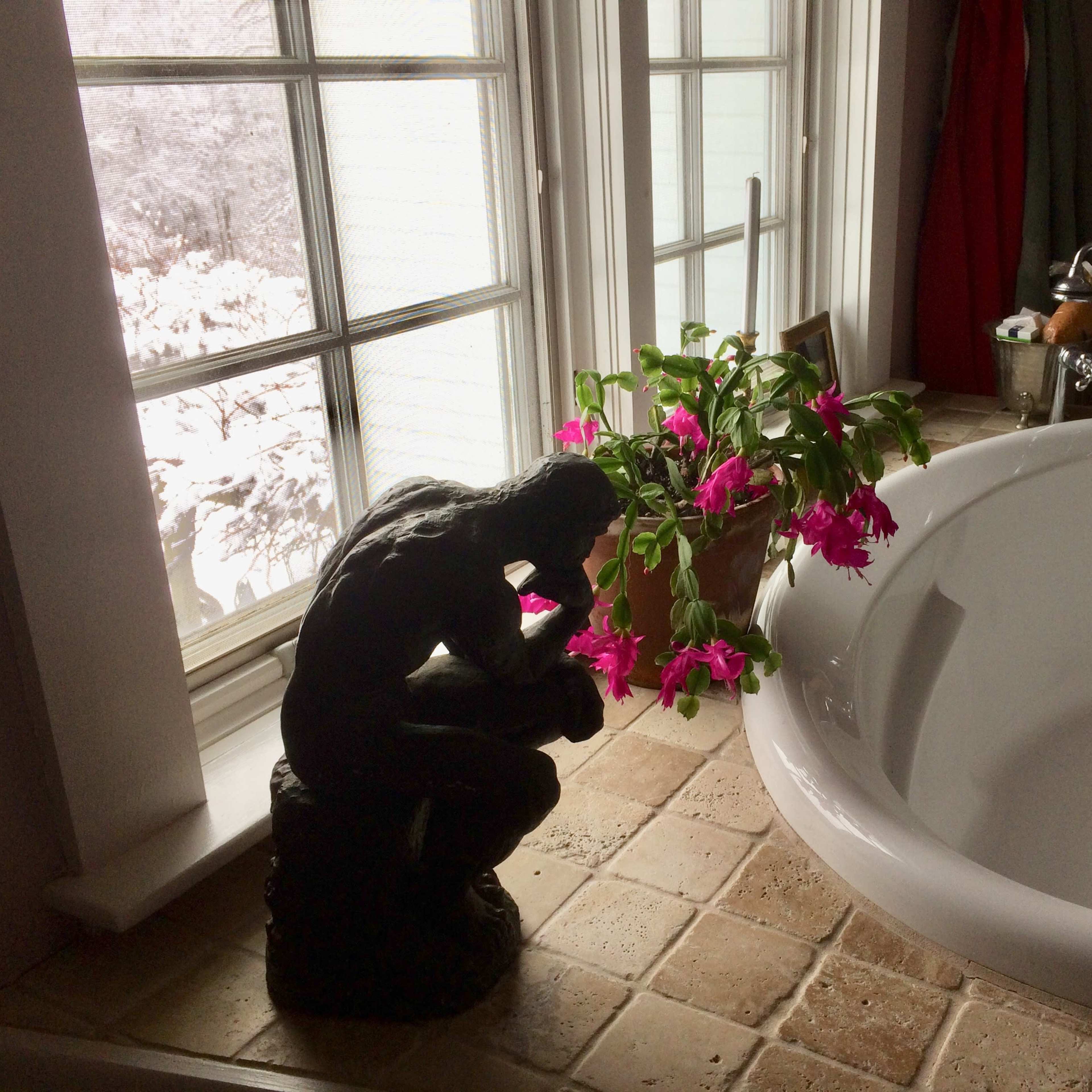 A sculpture of a contemplative figure sits next to a potted plant with pink flowers beside a freestanding bathtub in a well-lit bathroom.