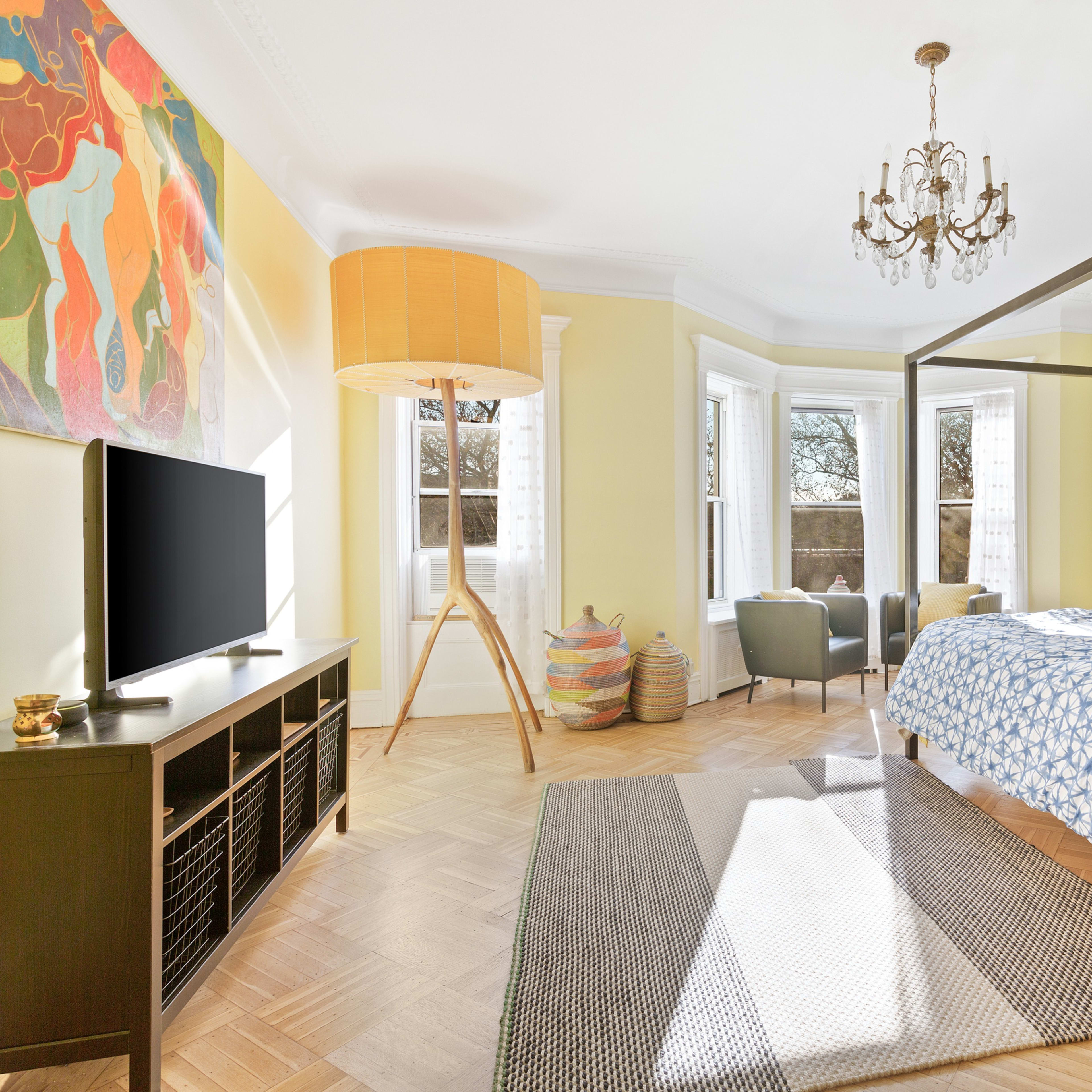 The scene depicts a bright bedroom featuring a four-poster bed with a patterned bedspread, a television on a black console, a large floor lamp, and windows that allow natural light.