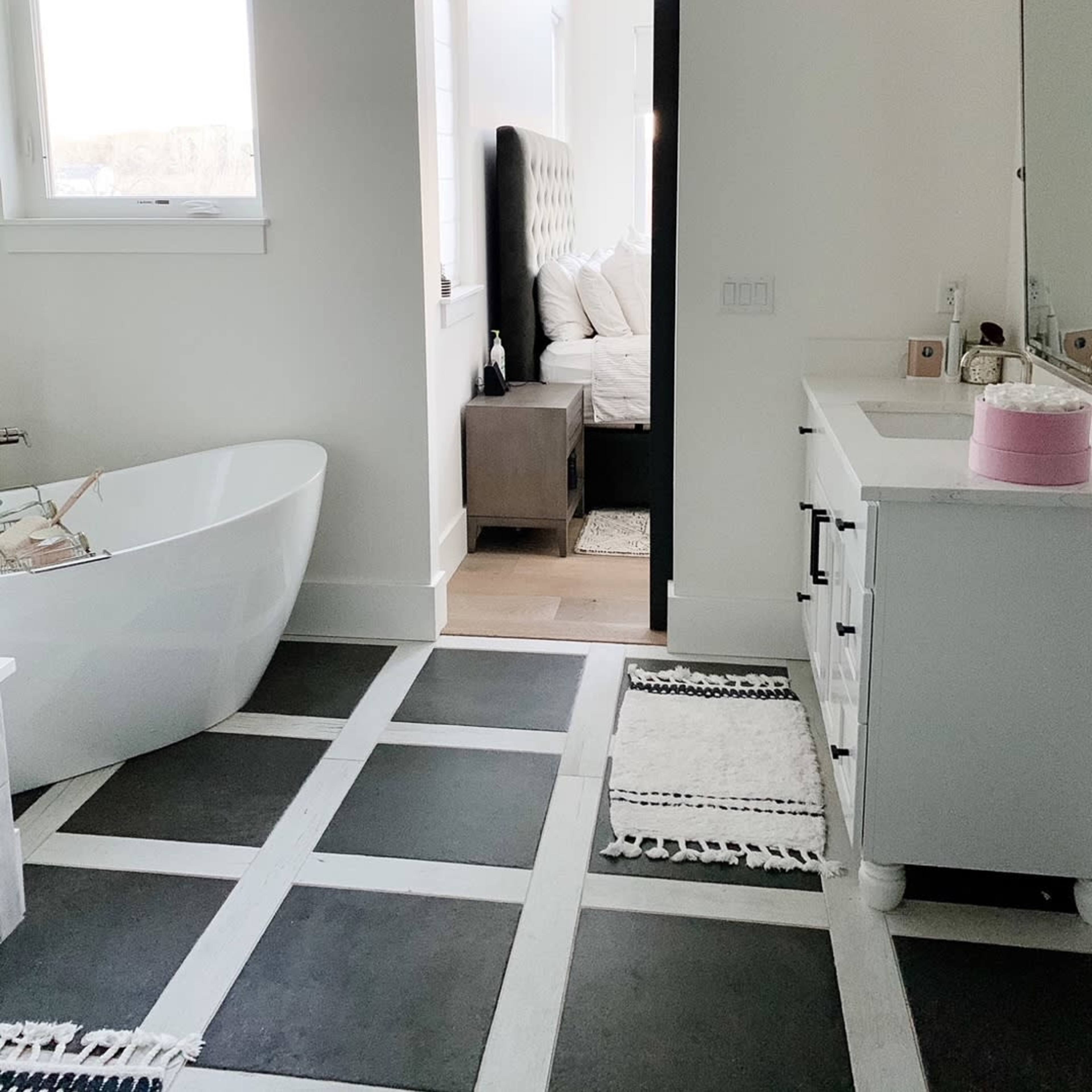 A bathroom with a freestanding bathtub, a doorway leading to a bedroom, and a vanity with a sink, all set on a tiled floor with black and white patterns.