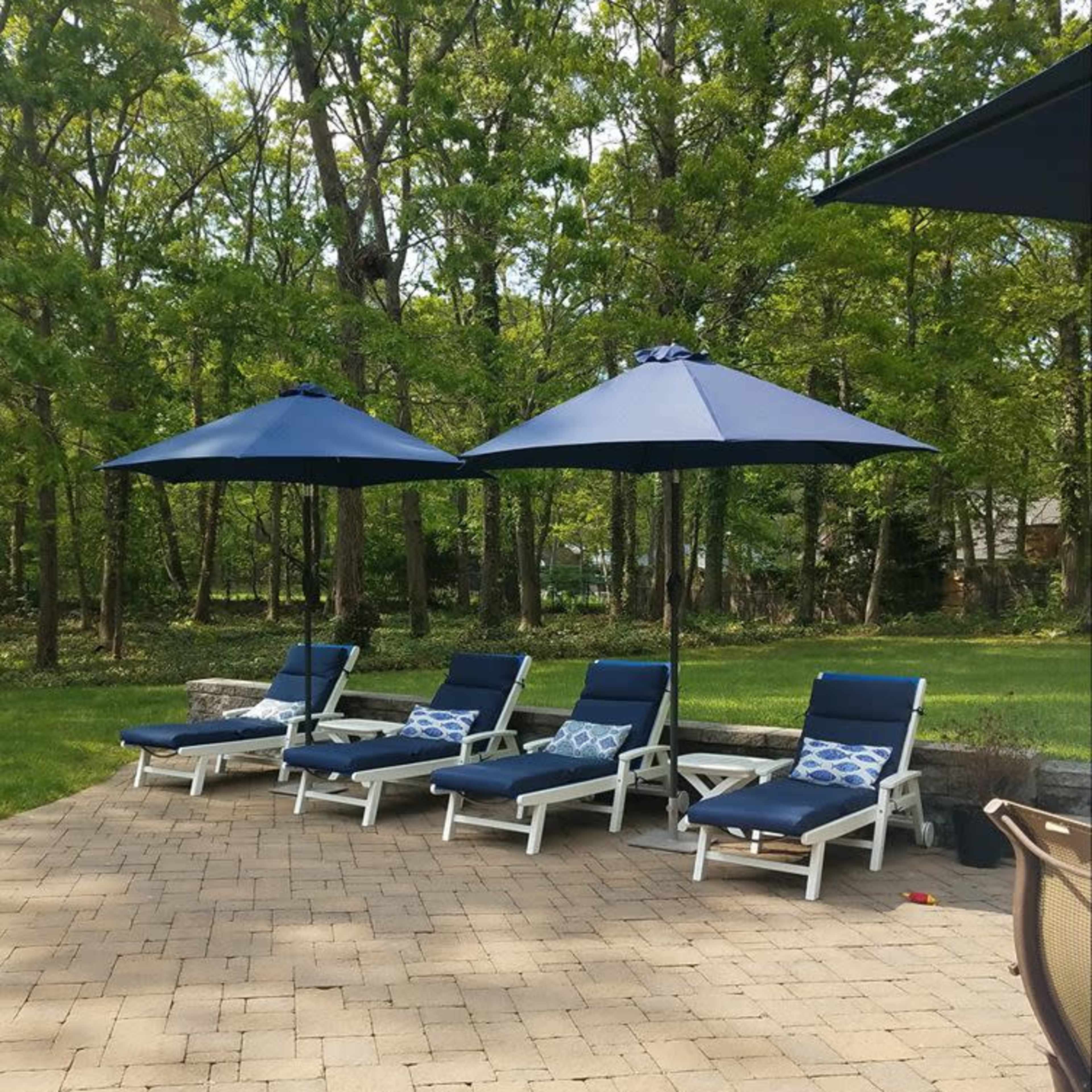 Three lounge chairs with blue cushions are arranged under navy umbrellas on a patio surrounded by trees.