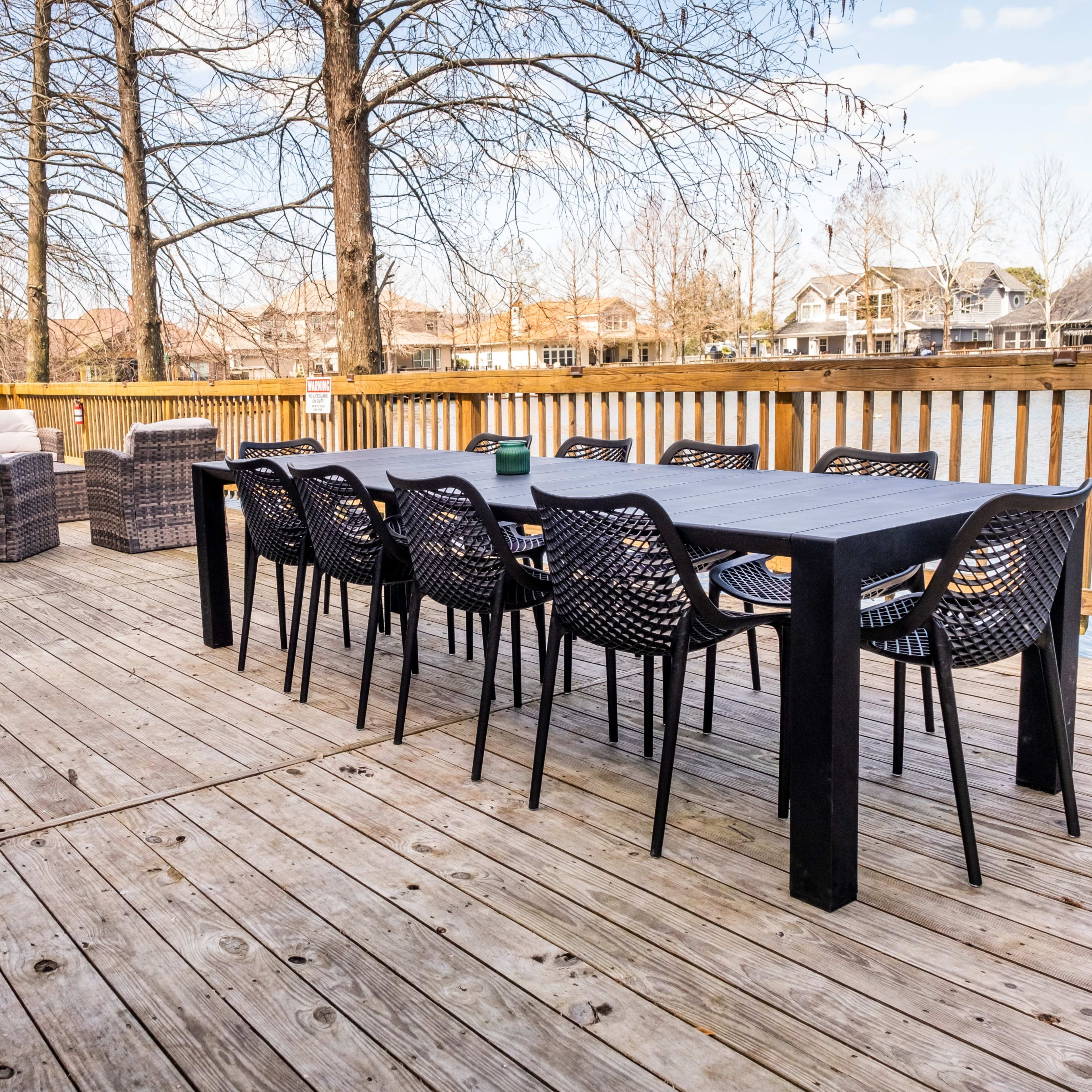 A long black table surrounded by matching chairs is set on a wooden deck near a lake, with wicker seating in the background.