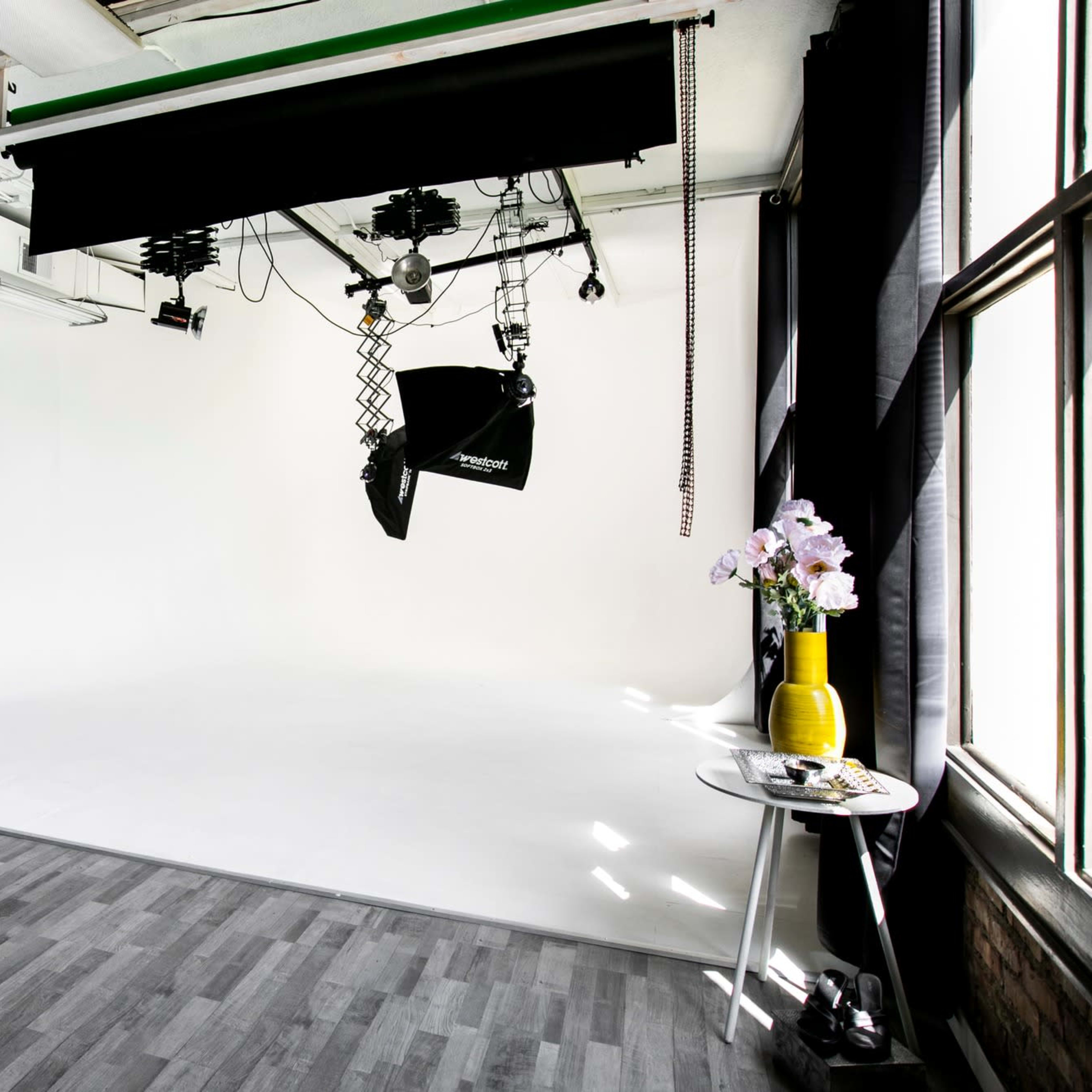 A photography studio features a white backdrop, large softbox lighting, wood flooring, and a mirror alongside decorative plants and a small table with flowers.