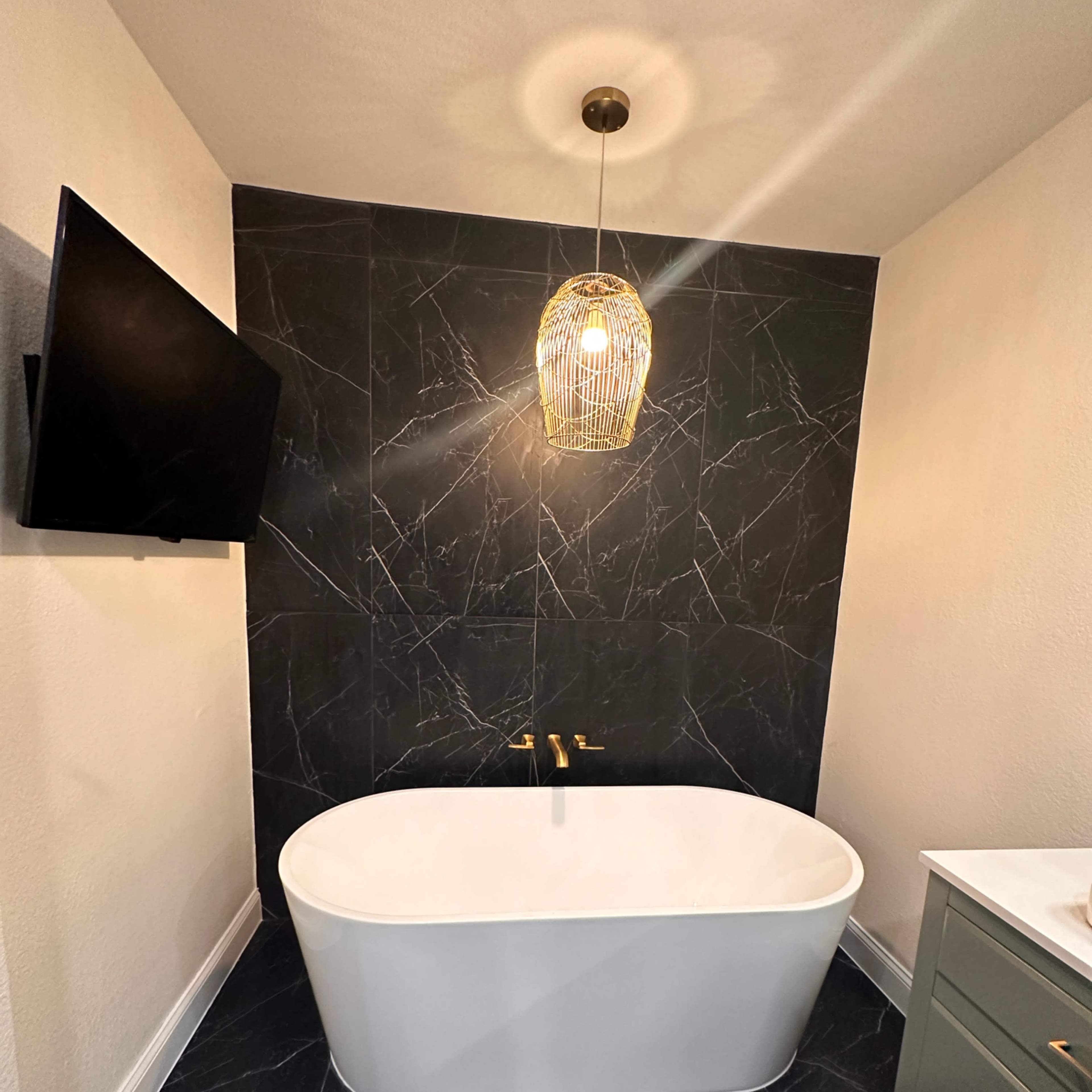 A modern bathroom featuring a freestanding white bathtub, a wall-mounted television, a pendant light, and dark marble walls.