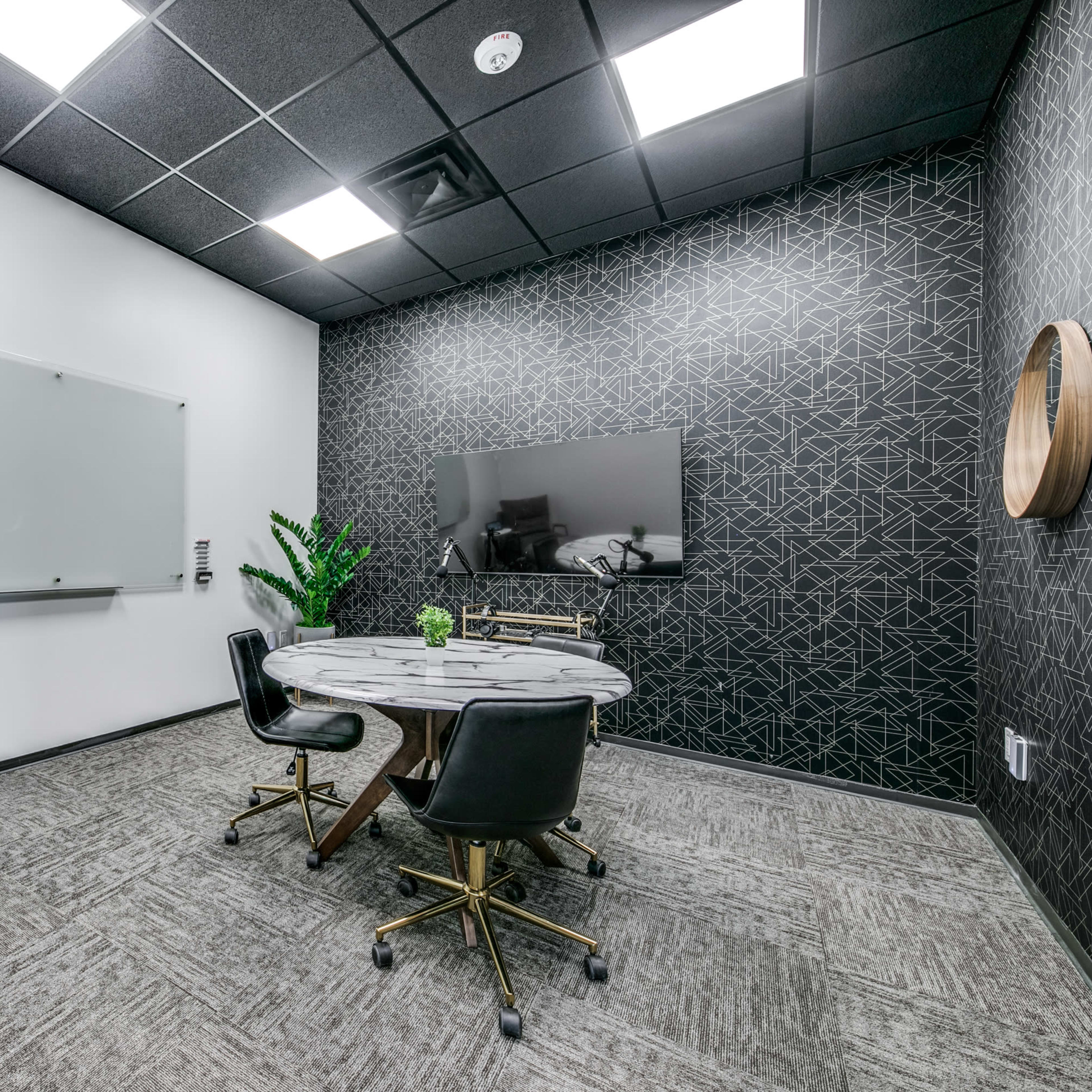 A small meeting room featuring a round table surrounded by four black chairs, a gray lounge chair, a whiteboard, and a TV on a patterned wall.
