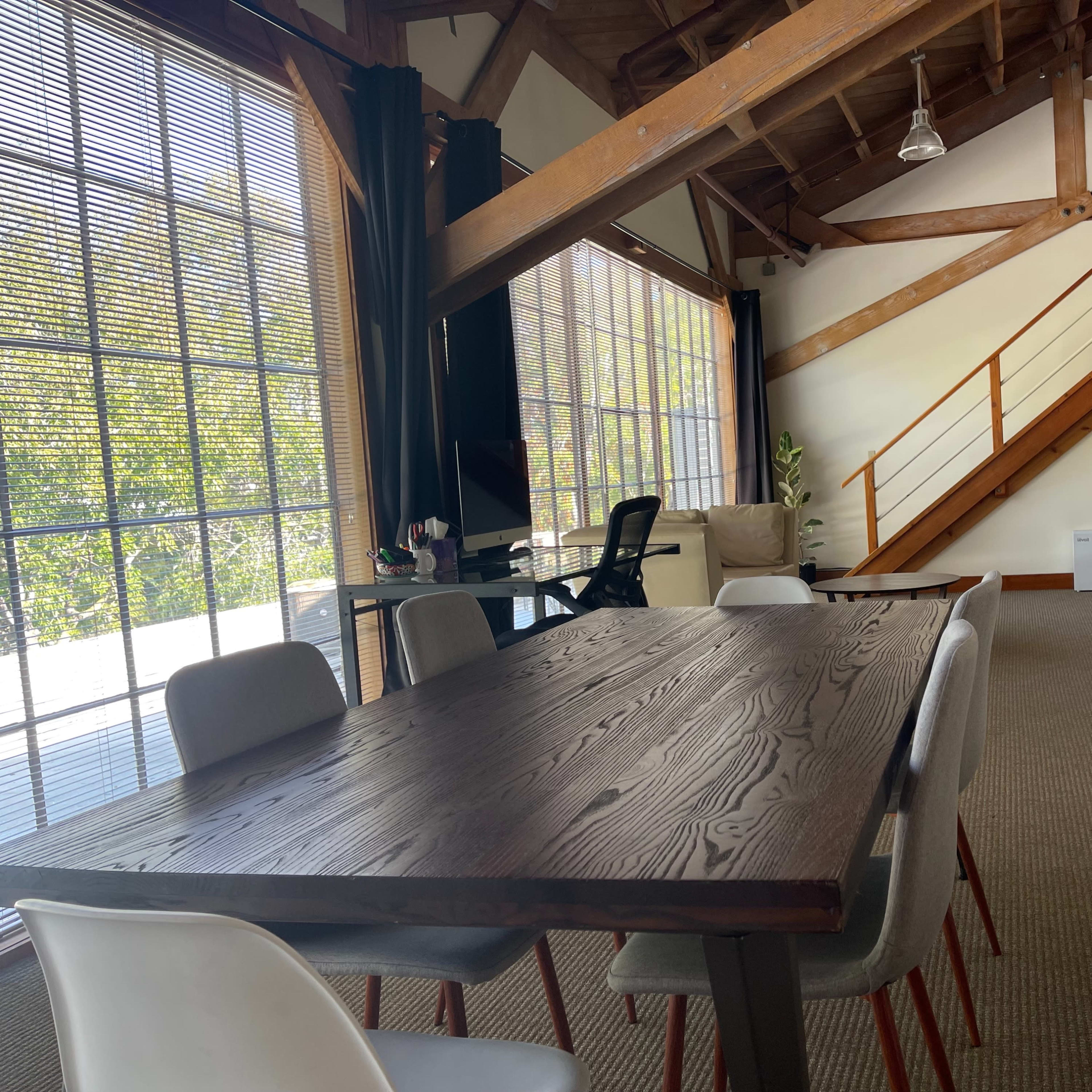 A spacious interior with a wooden table surrounded by chairs, large windows letting in natural light, and an upper staircase leading to a loft area.