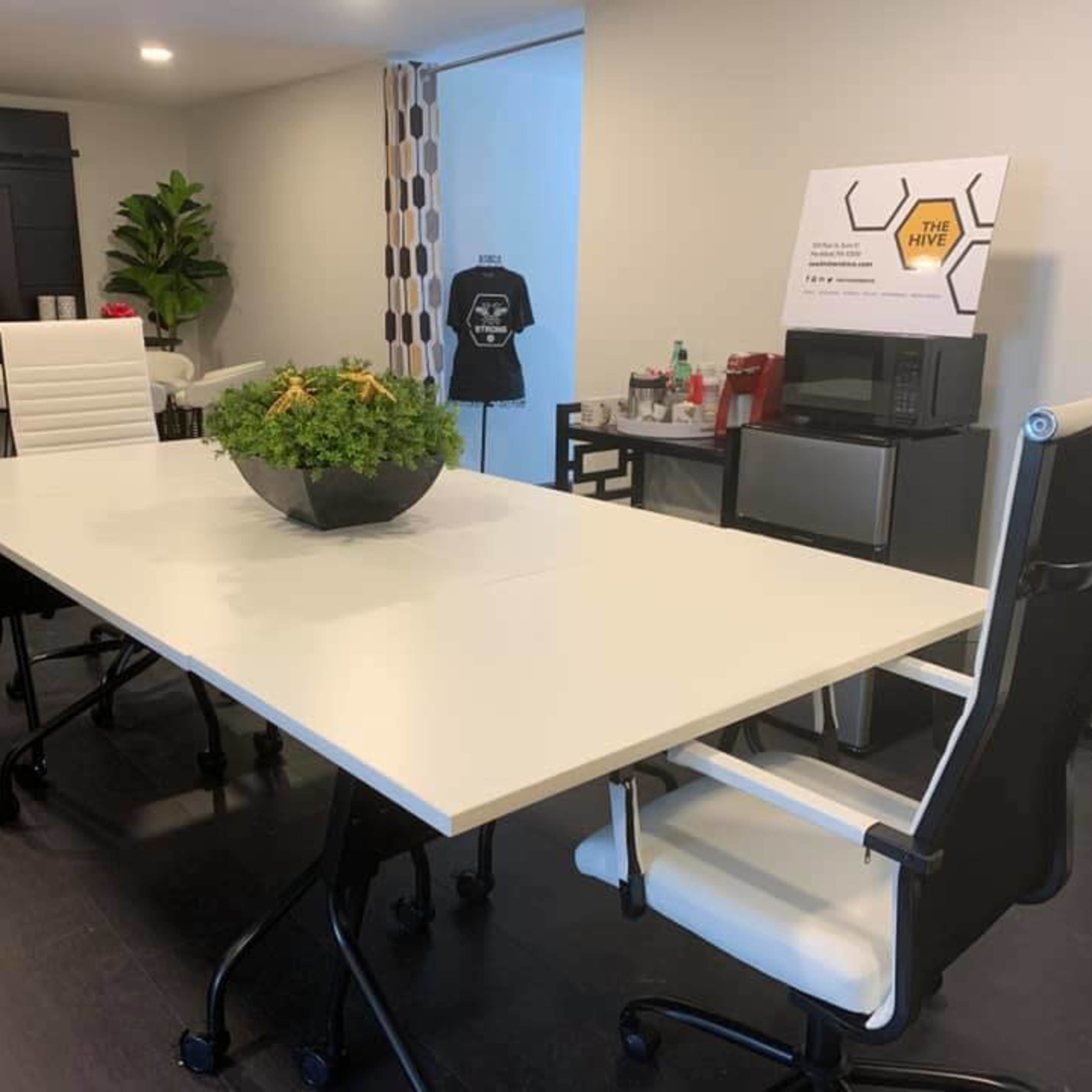 A modern conference room features a large white table surrounded by black and white chairs, with a potted plant centerpiece and a sign displaying "THE HIVE."