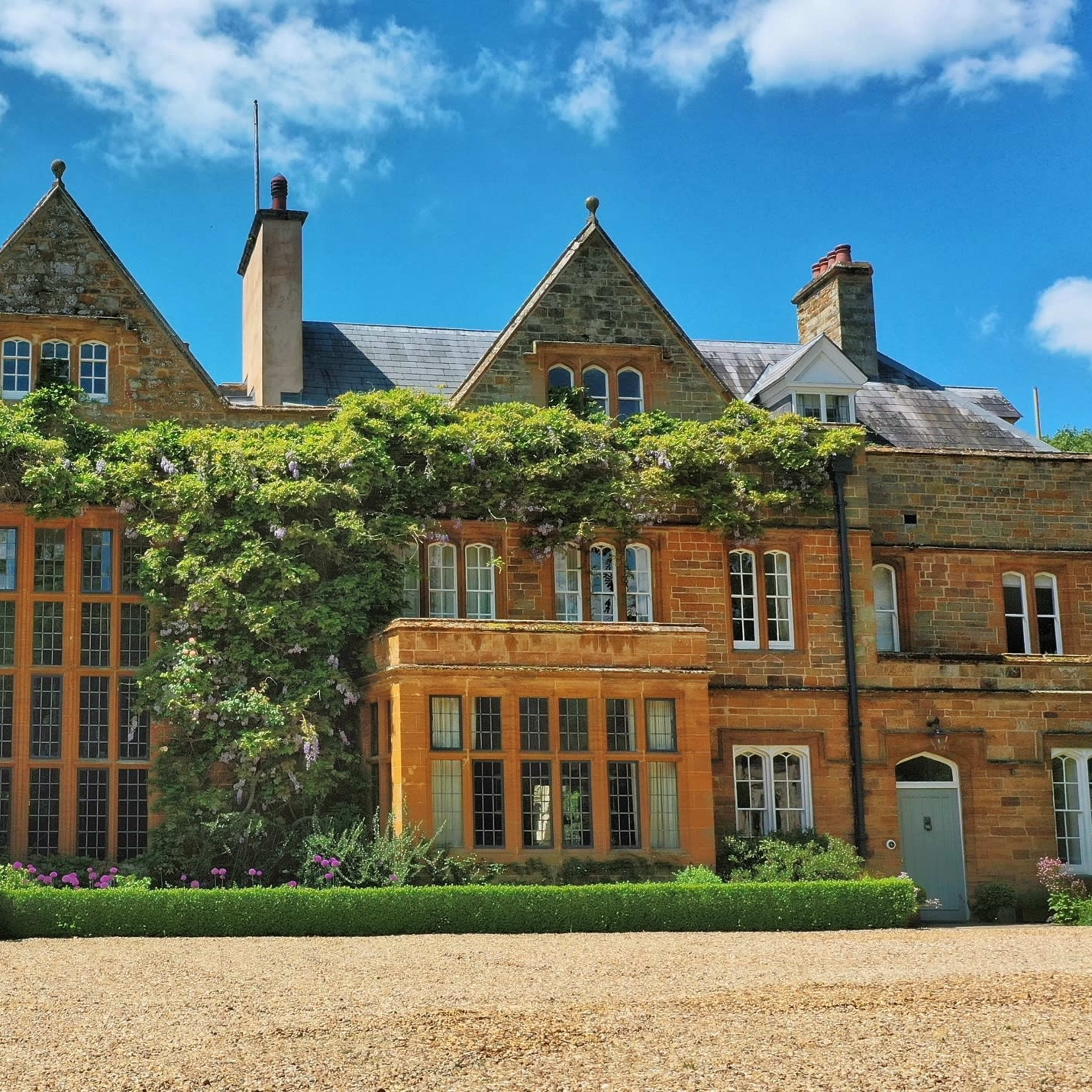 A large, brick mansion with multiple gabled roofs, adorned with greenery and surrounded by a neatly landscaped garden.
