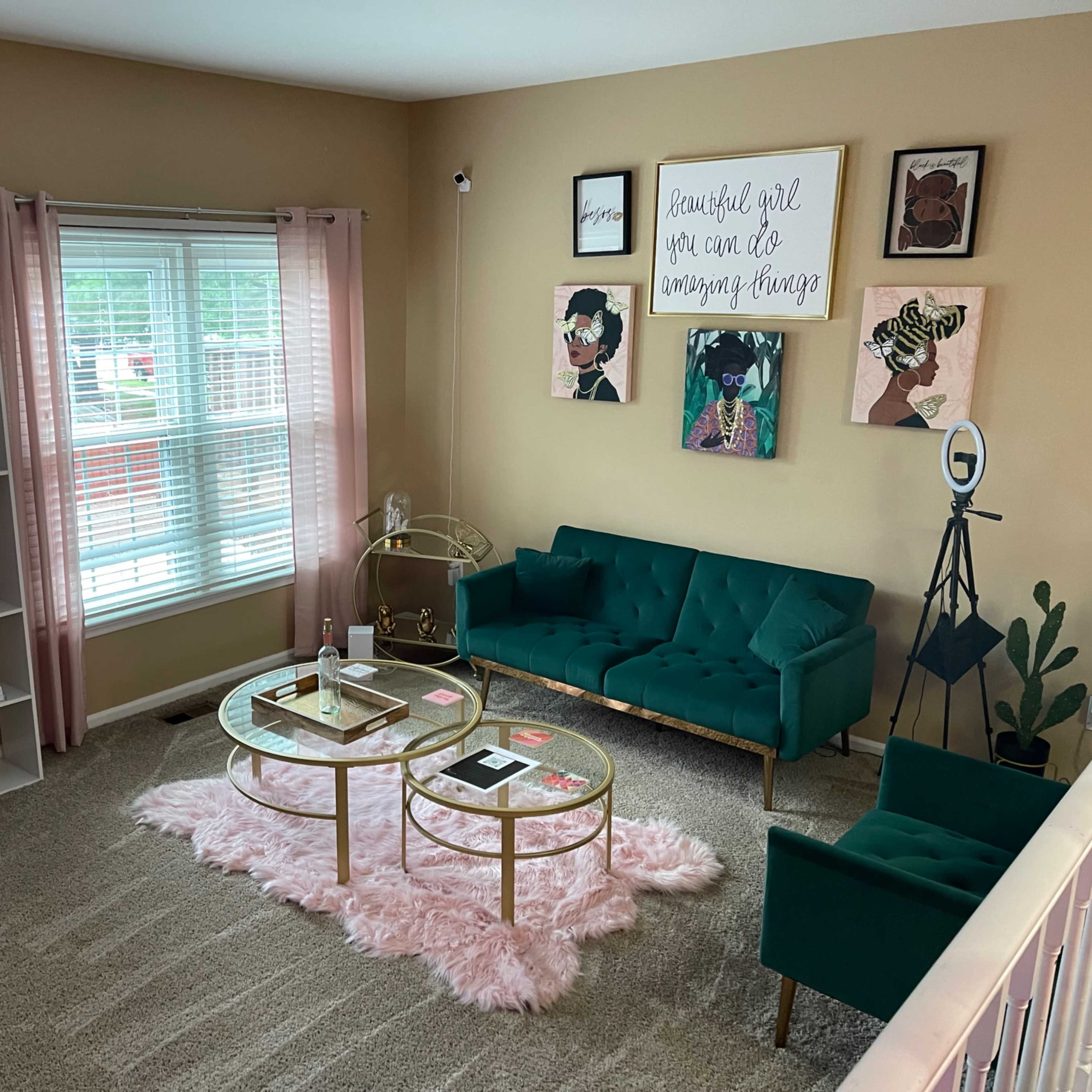 A living room with a green couch, two gold coffee tables on a pink rug, and a bookshelf filled with decorative items against a wall adorned with framed artwork.