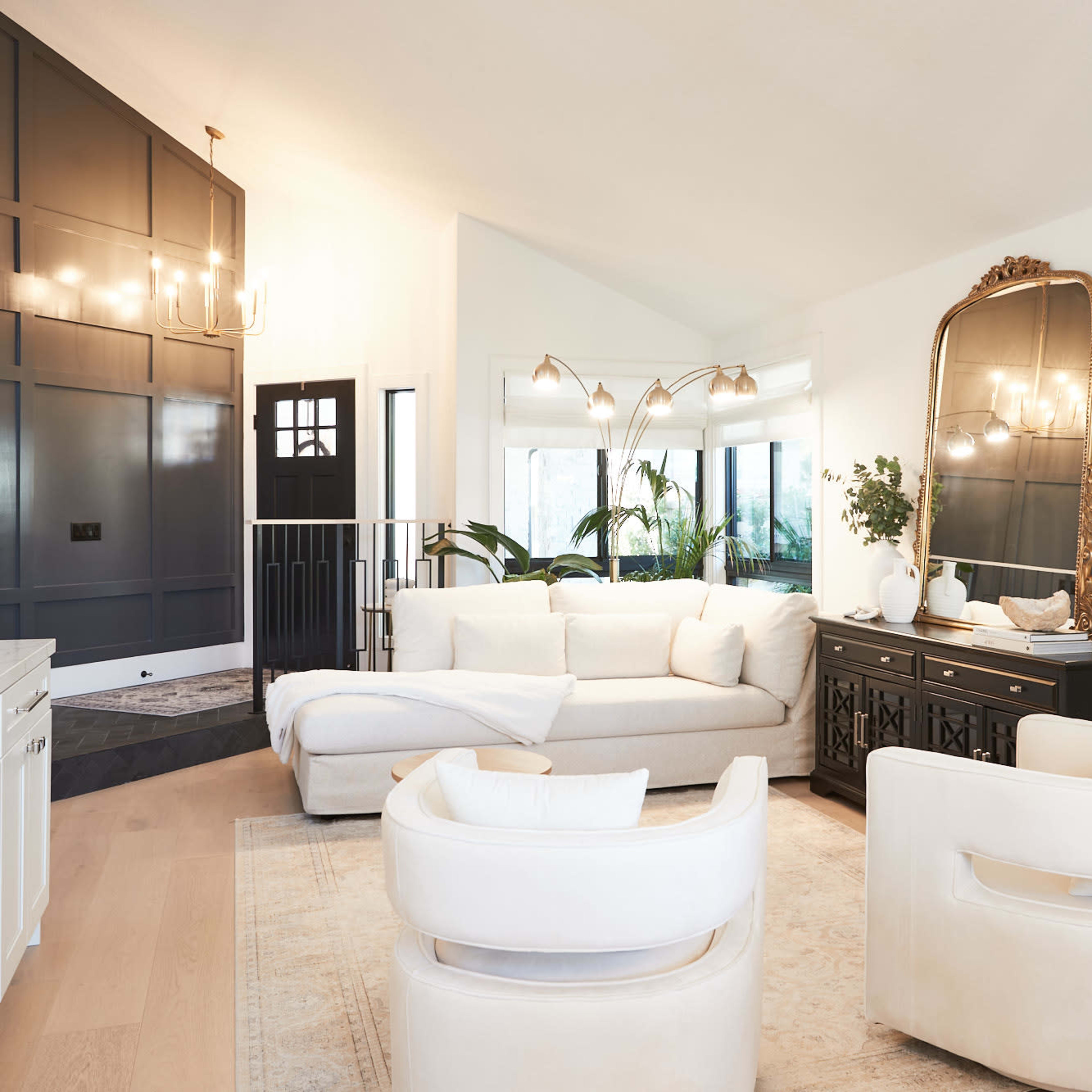 A modern living room featuring a white sectional sofa, two white armchairs, a black and gold mirror, and large windows overlooking greenery.