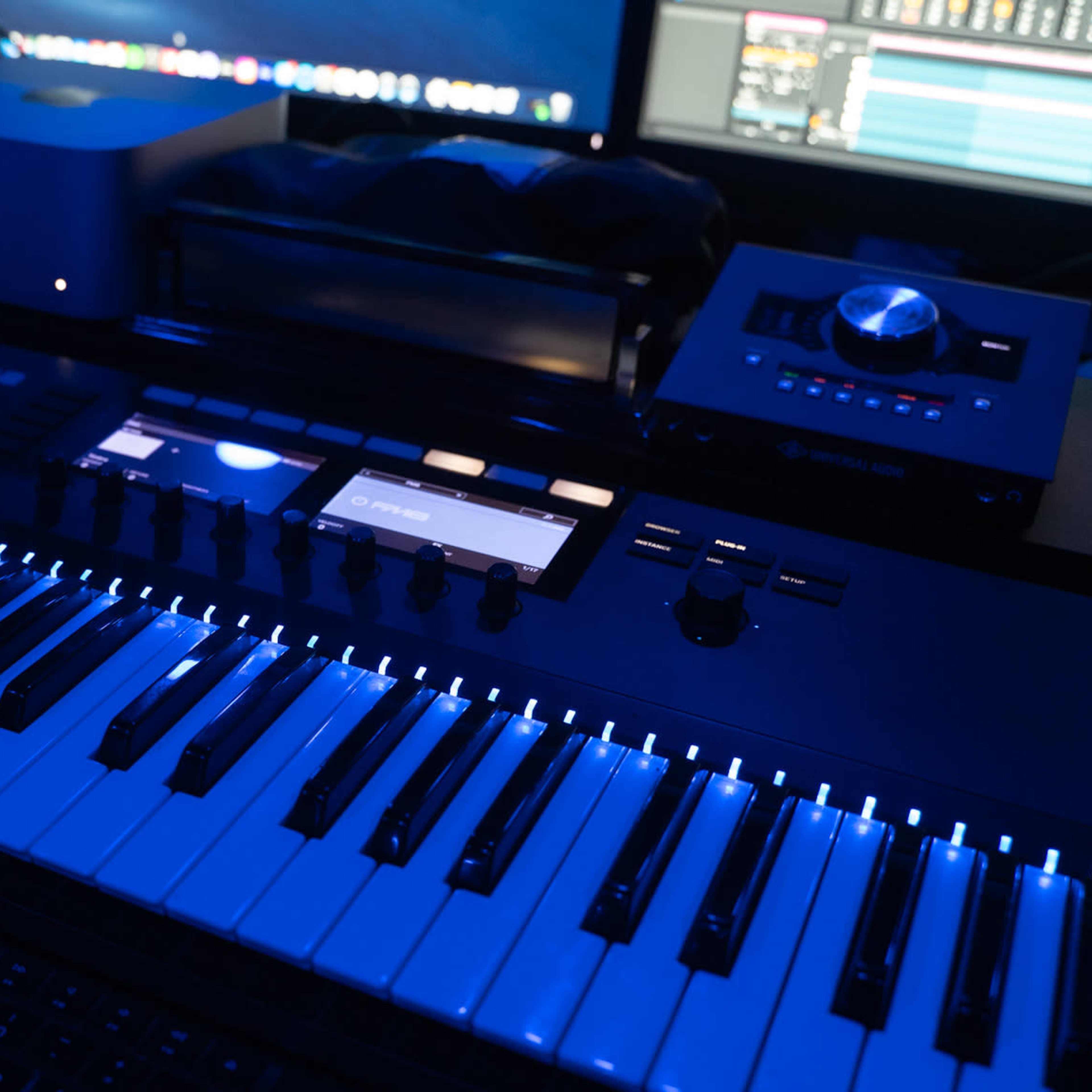 A close-up of a black MIDI keyboard with illuminated keys and control buttons, situated in a music production studio with computer screens in the background.