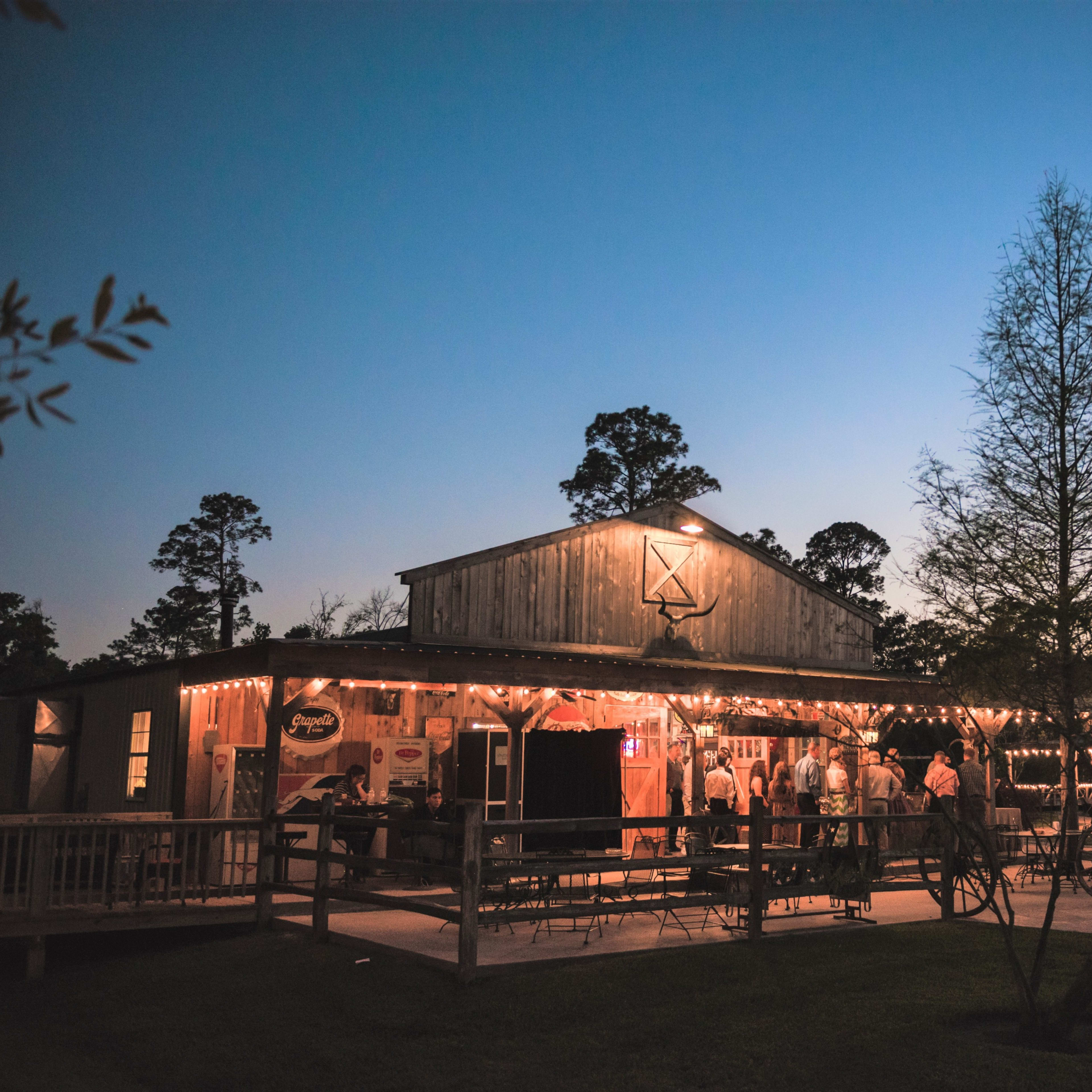 A rustic venue with string lights and a wooden exterior is bustling with people as twilight sets in.