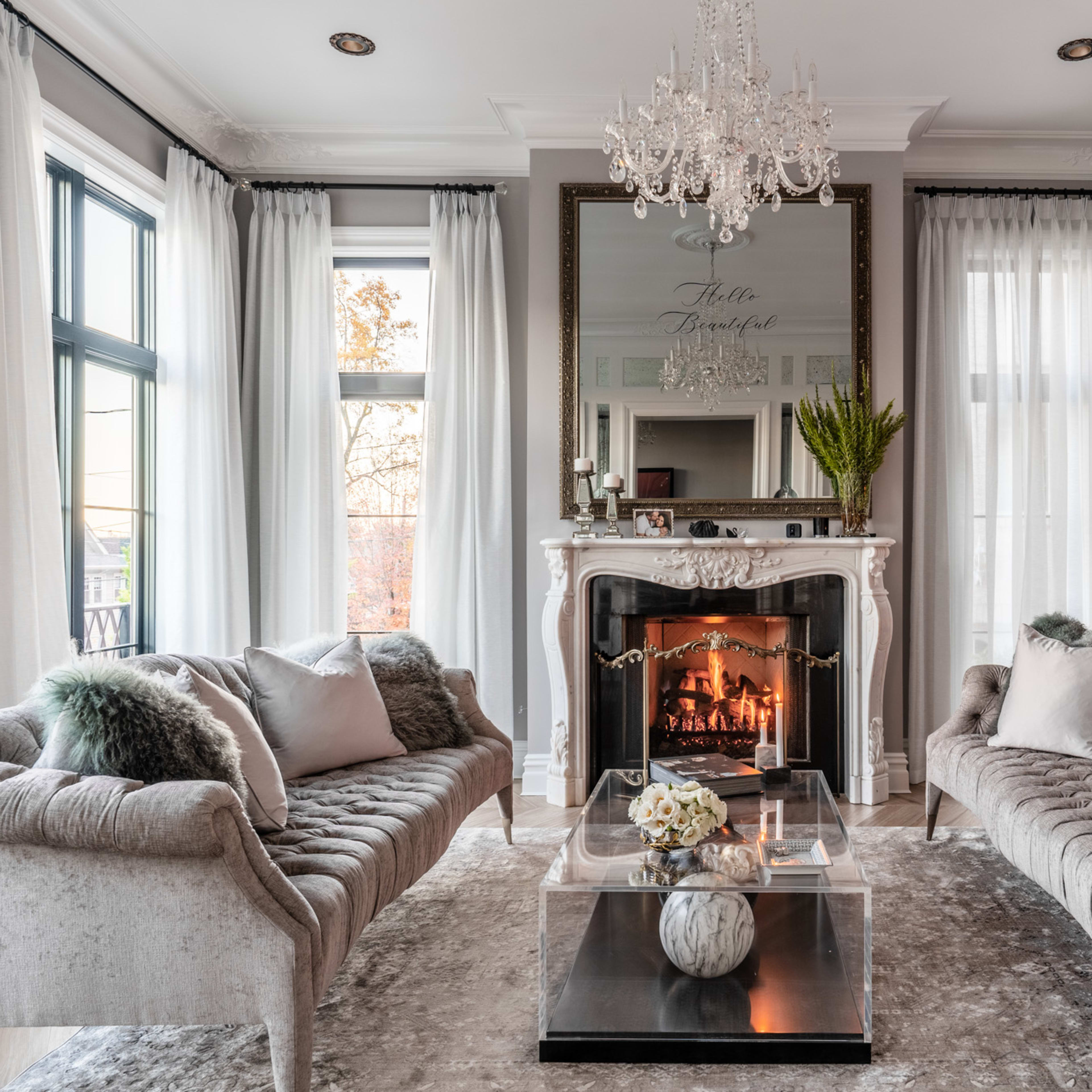 The living room features two elegant sofas facing a decorative fireplace, with a large mirror and a chandelier above, and large windows providing natural light.