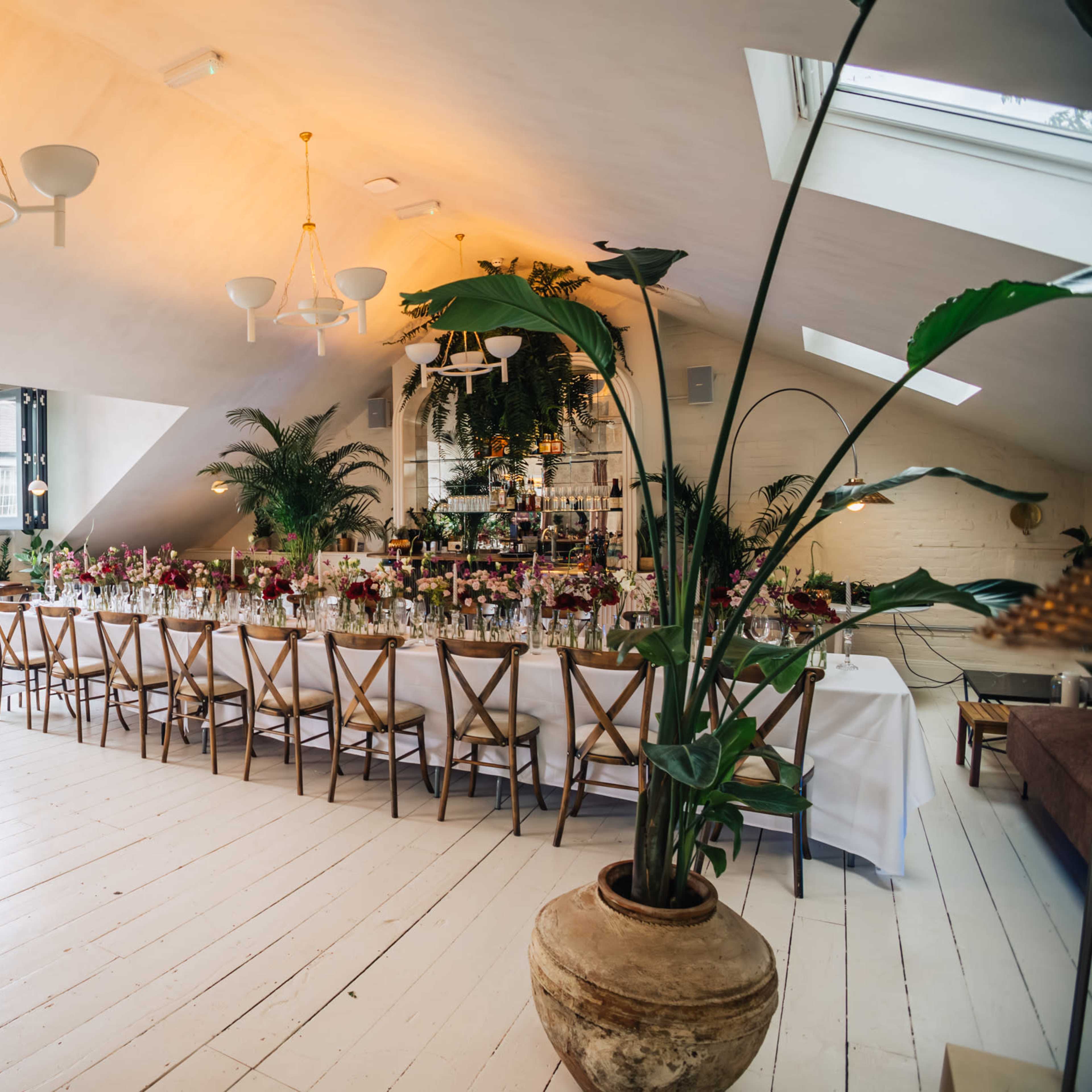 A bright, spacious event room features a long table set for a gathering, surrounded by greenery and modern decor under a sloped ceiling.
