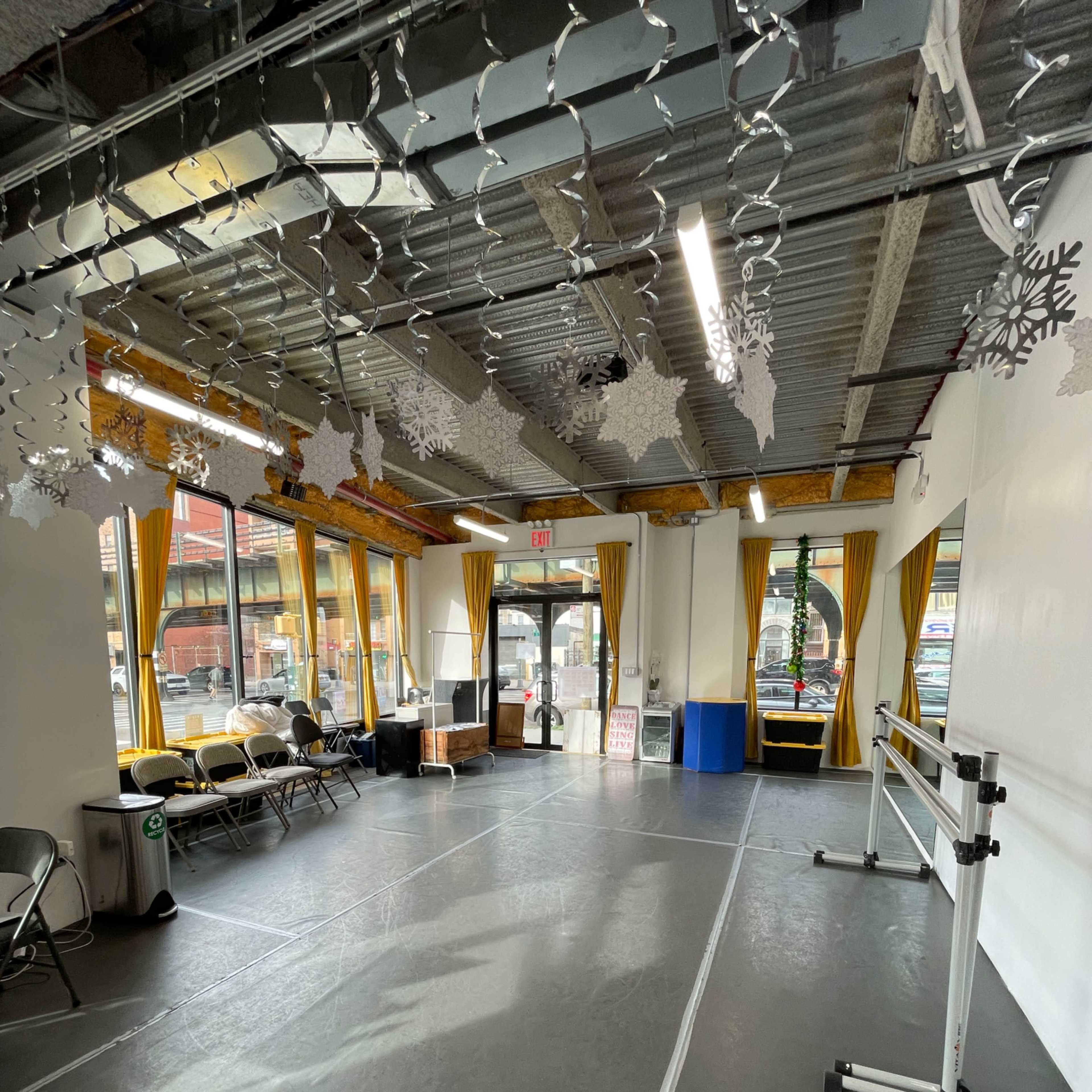 A bright room with a gray floor, large windows, and chairs arranged along the walls, featuring ballet barres on one side and winter decorations hanging from the ceiling.