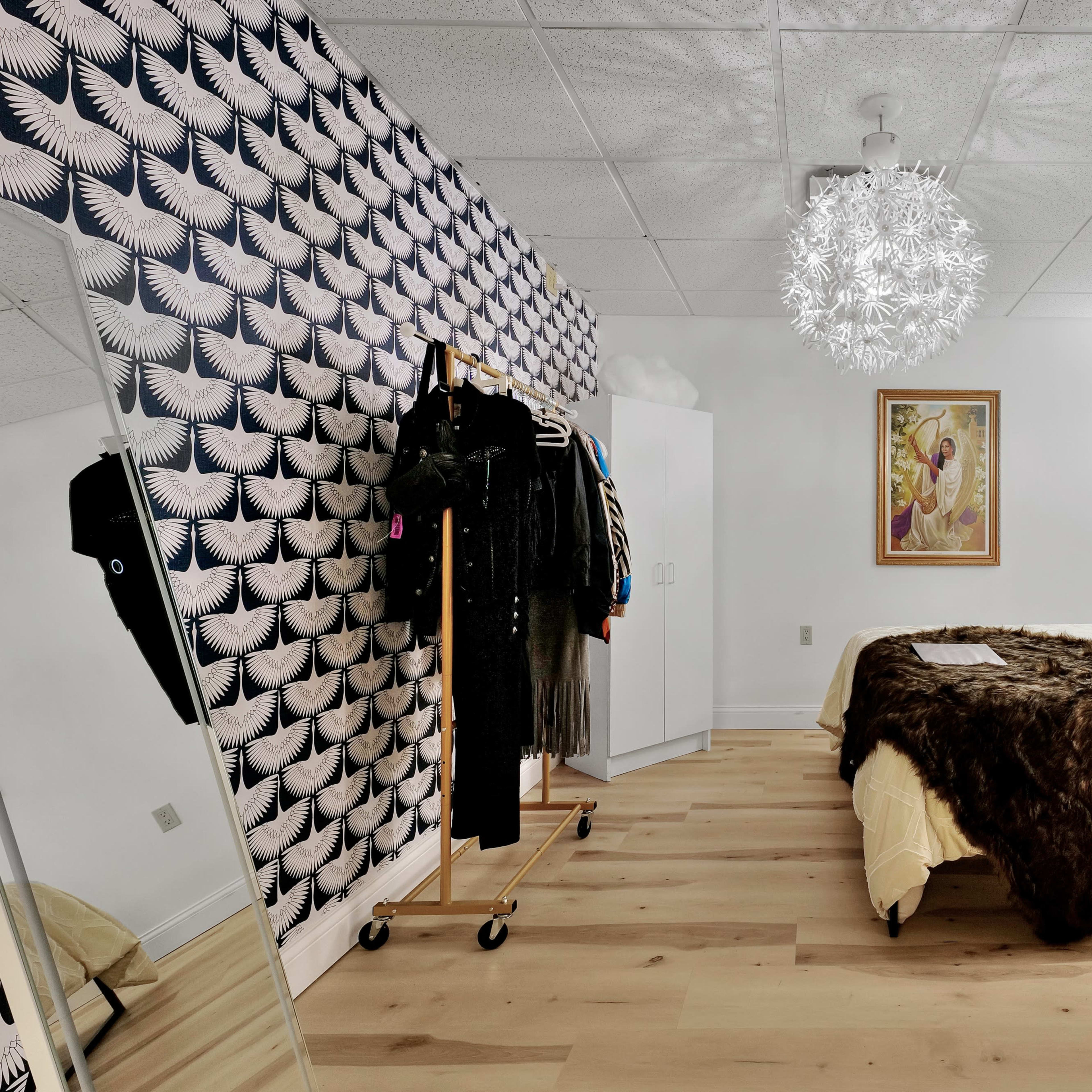 The room features a bed with a brown blanket, a mirrored wardrobe, and a patterned wall with hanging clothing on a rack.