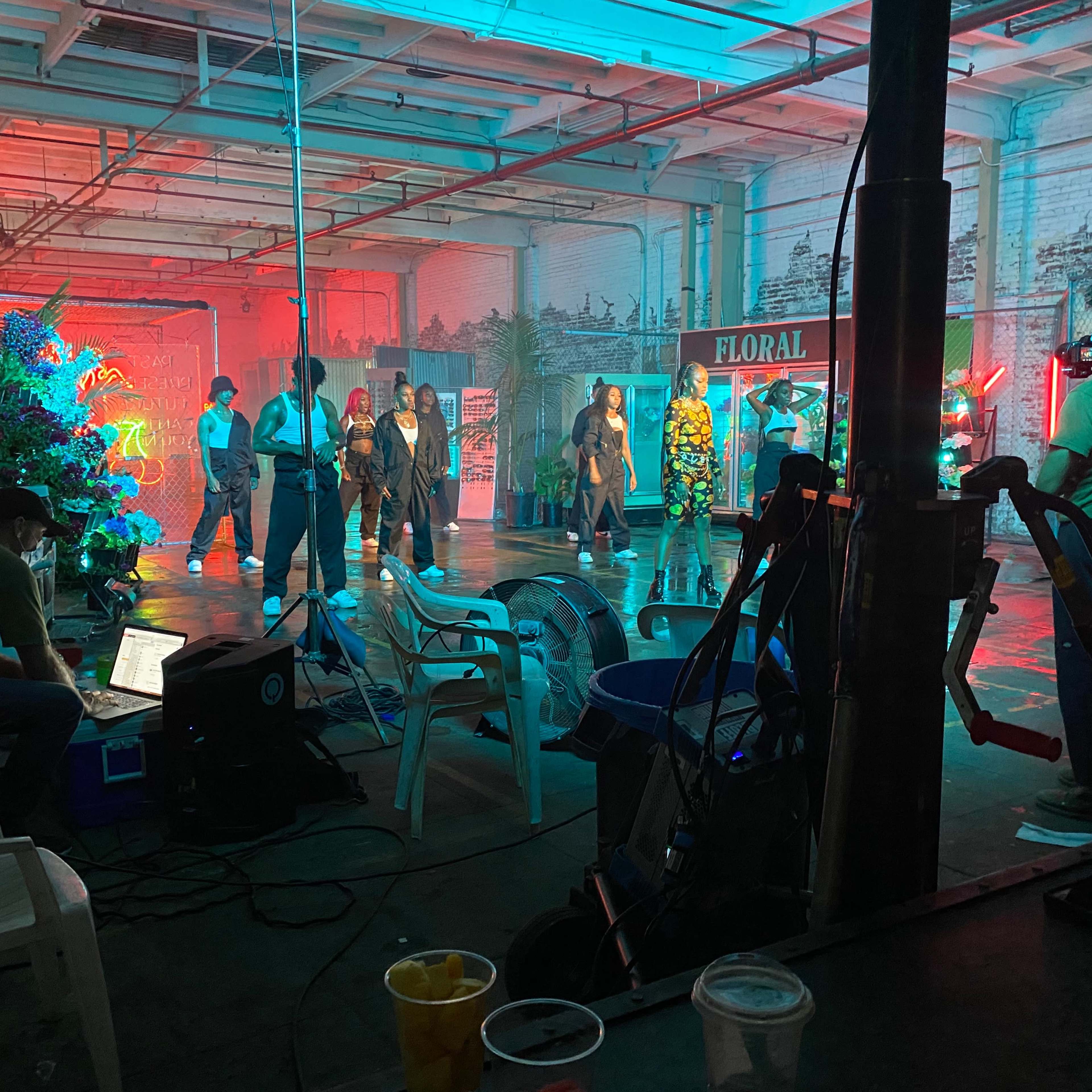 A group of performers rehearses on a brightly lit stage inside an industrial building, while crew members manage equipment and monitor the scene from the sidelines.