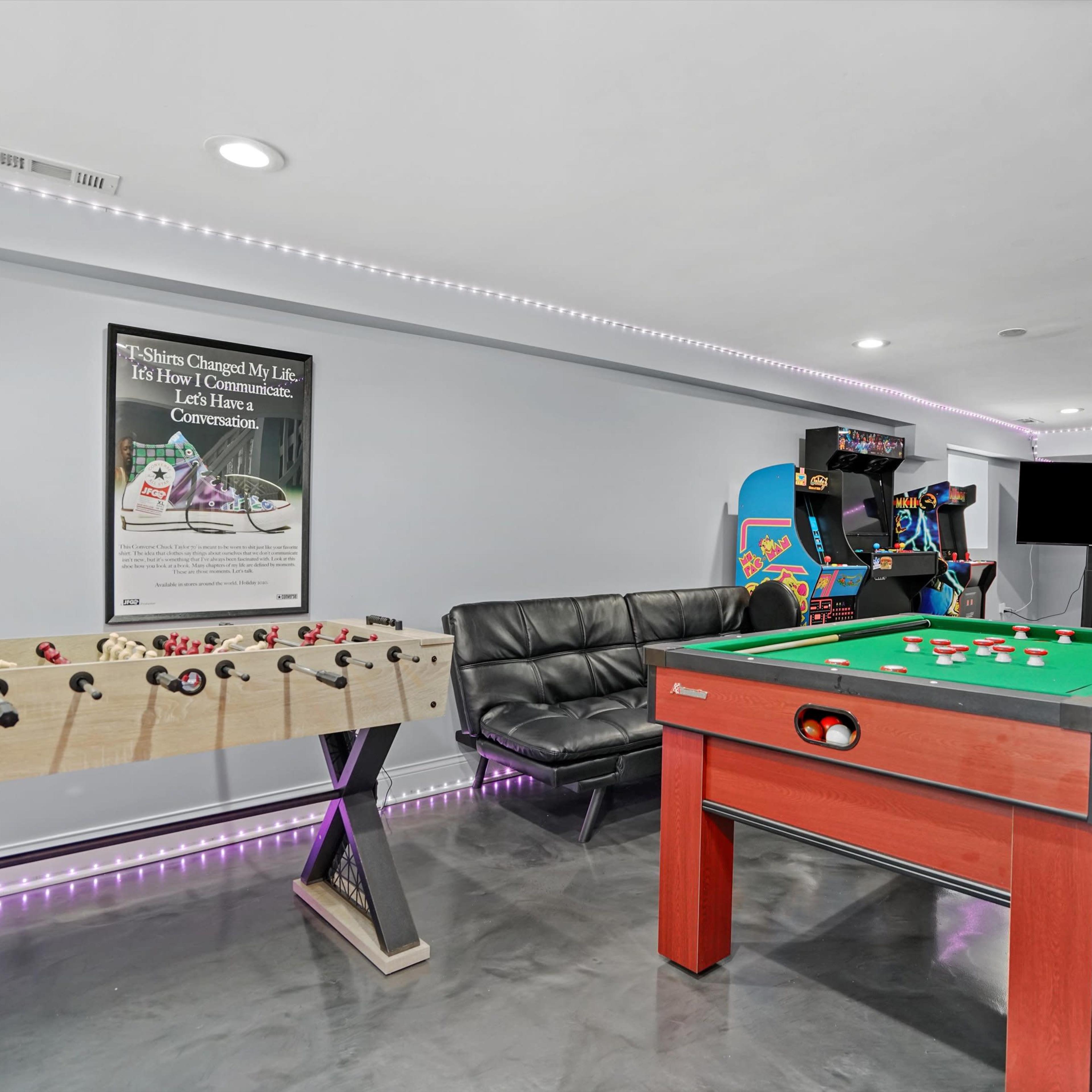 A modern game room featuring a foosball table, a pool table, a black couch, and arcade games, with ambient lighting along the walls.