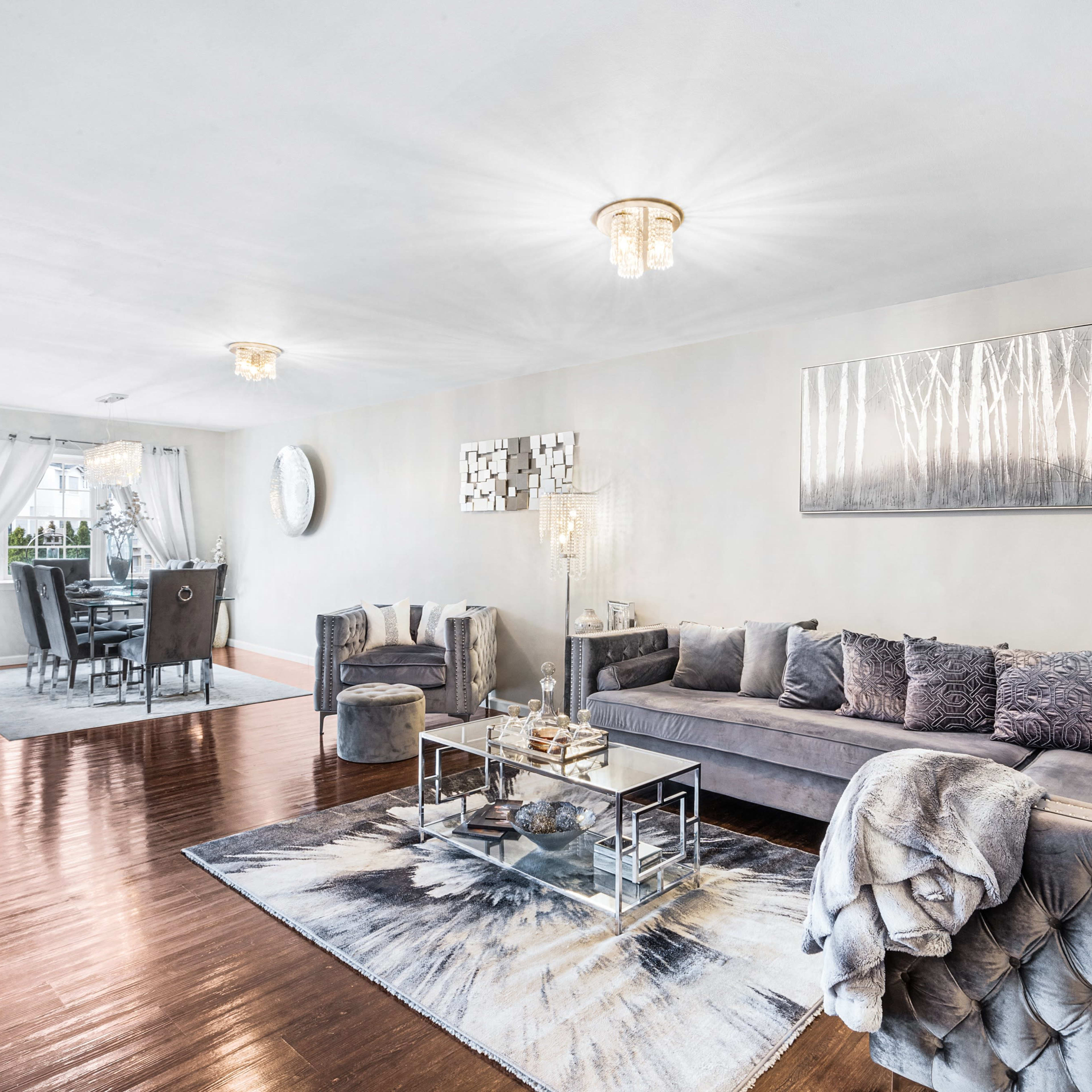 A modern living room with a gray sectional sofa, glass coffee table, and decorative artwork, opening to a dining area in the background.