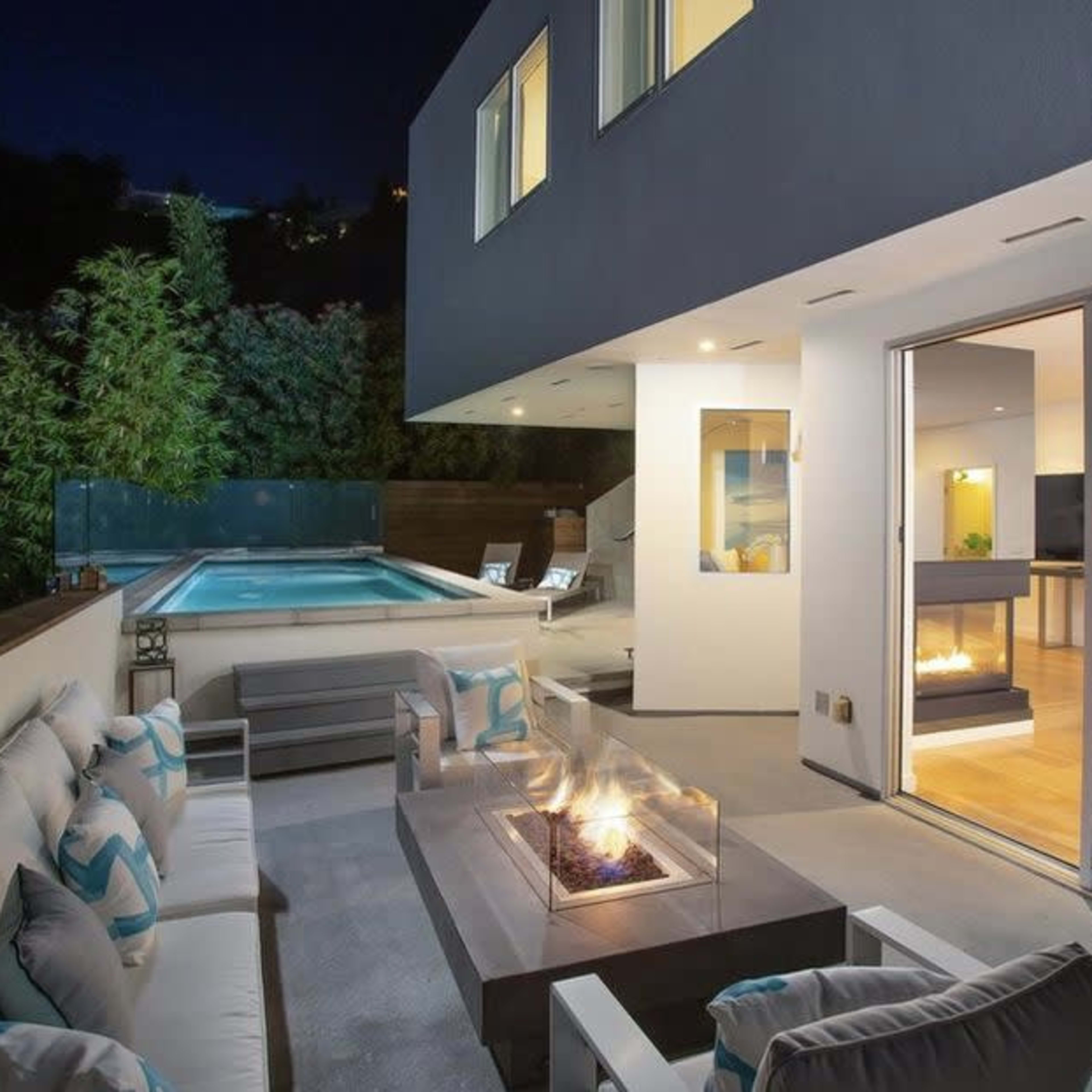 A modern outdoor seating area with a fire pit is adjacent to a swimming pool and a glass-walled interior space illuminated at night.