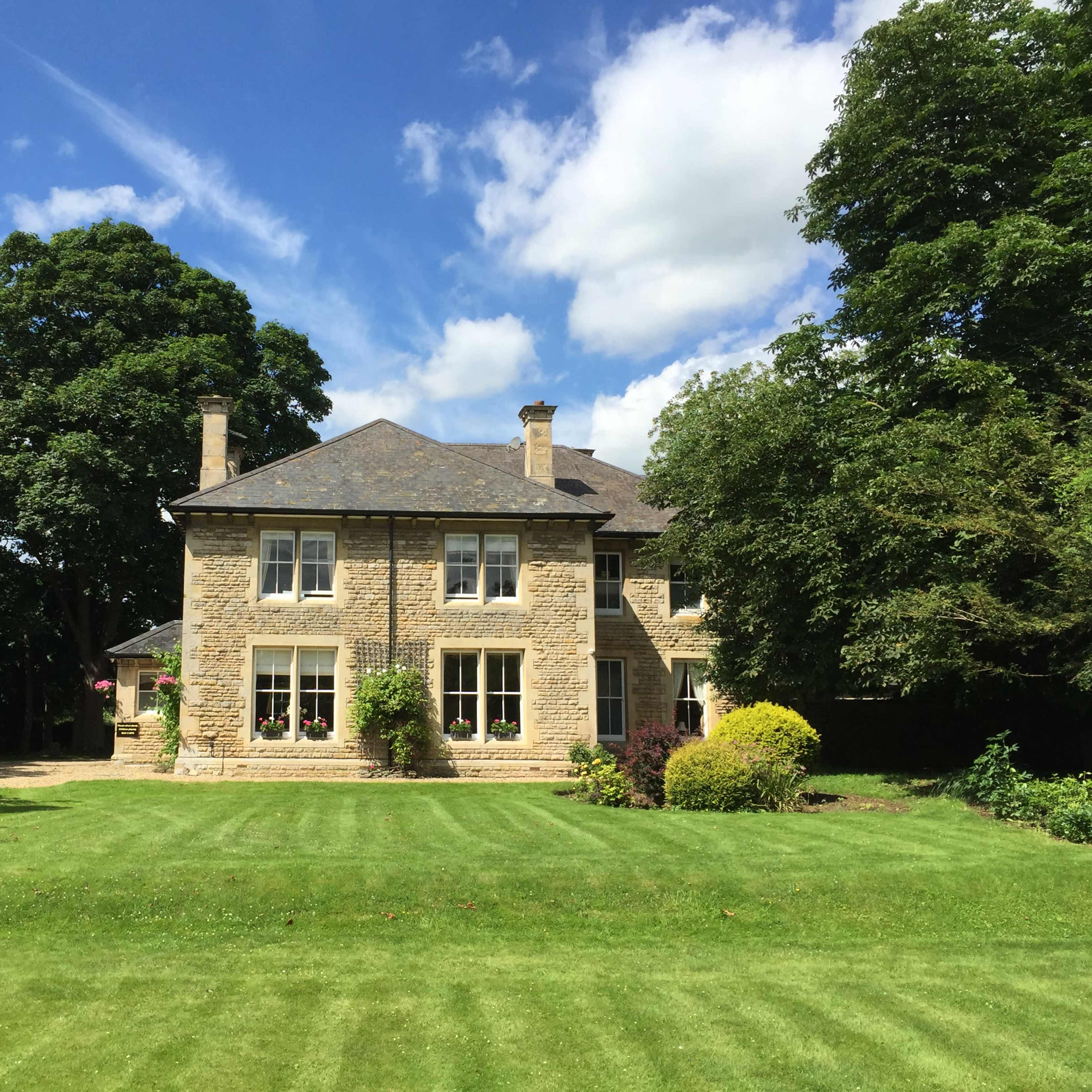 A large stone house with several windows is situated on a well-manicured lawn surrounded by trees under a partly cloudy sky.