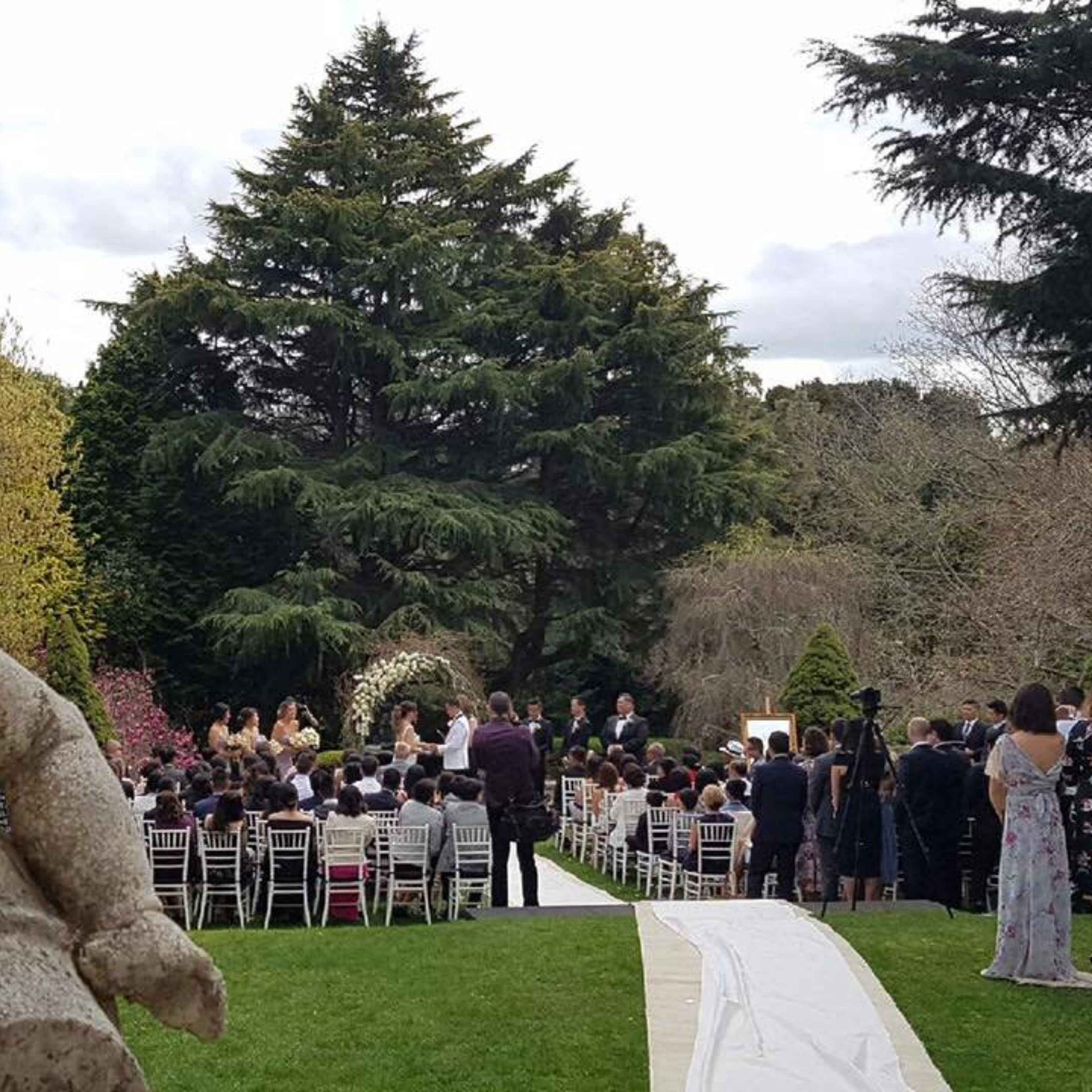 A wedding ceremony is taking place outdoors in a garden, with guests seated on white chairs and a floral arch in the background.