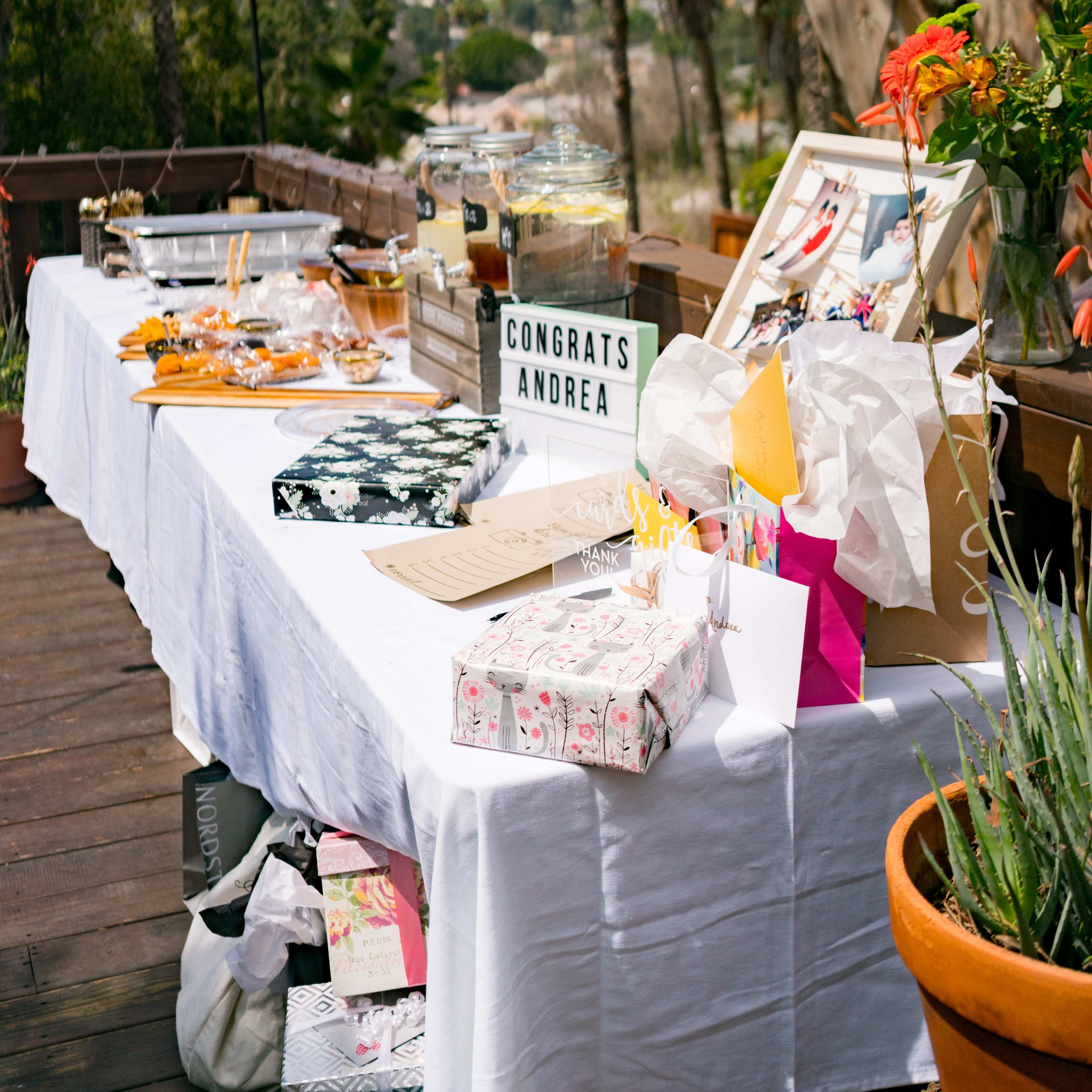 A table covered with a white cloth is adorned with gifts, snacks, and a sign that reads "CONGRATS ANDREA," set against a backdrop of palm trees and colorful decorations.