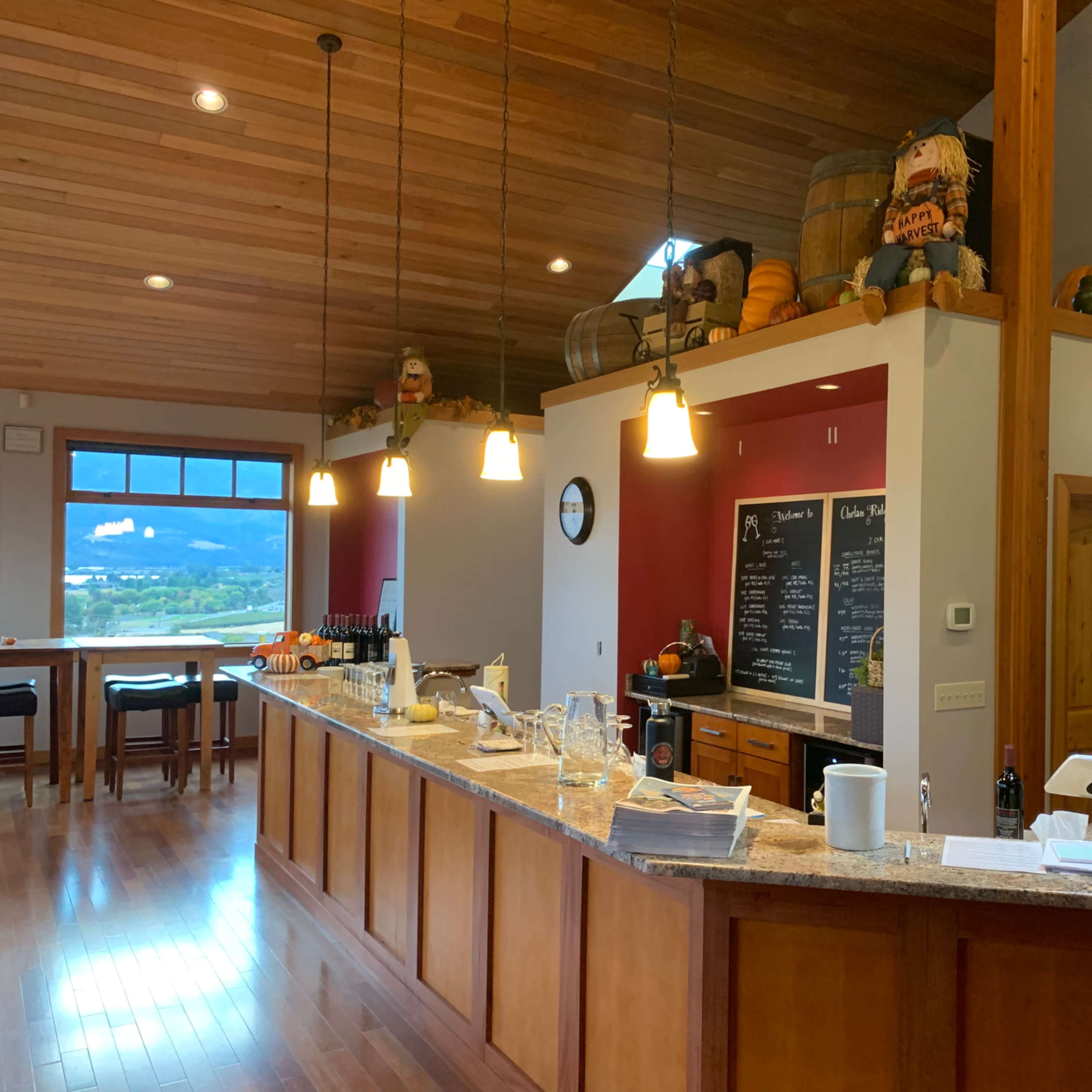 A spacious wine tasting room with a wooden bar, glassware, bottles of wine, and a chalkboard menu, surrounded by warm wood accents and decorative items.
