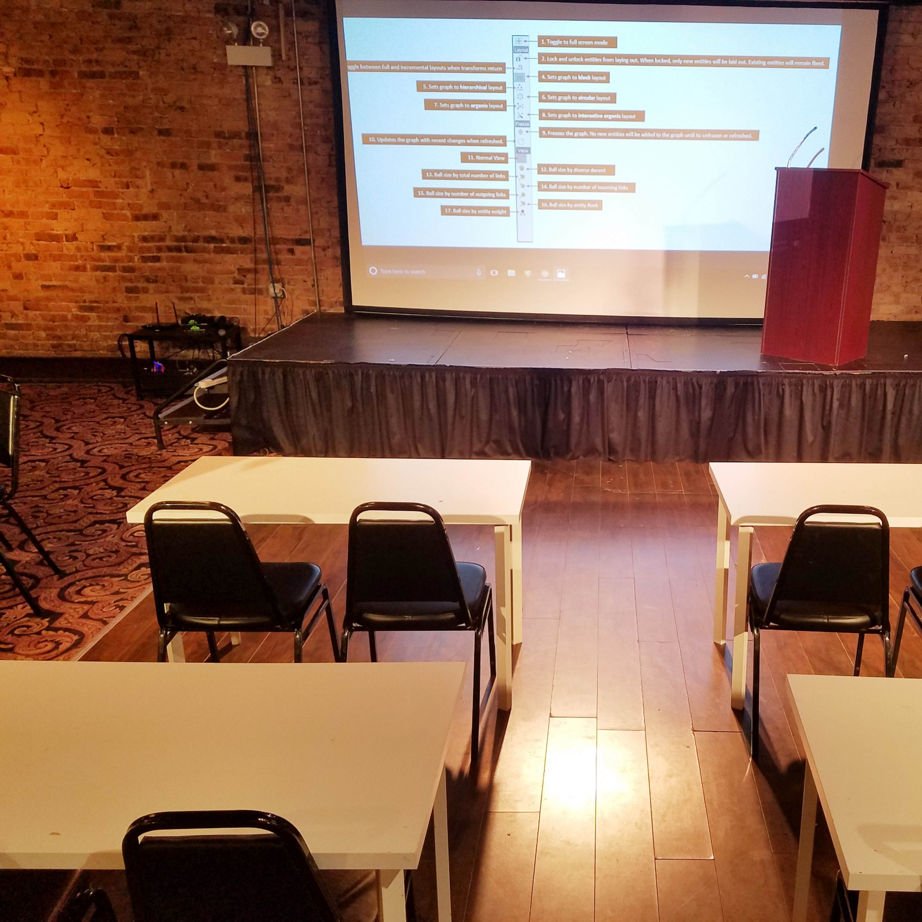 A presentation setup in a room with a stage, a podium, and several rows of empty tables and chairs facing a screen displaying content.