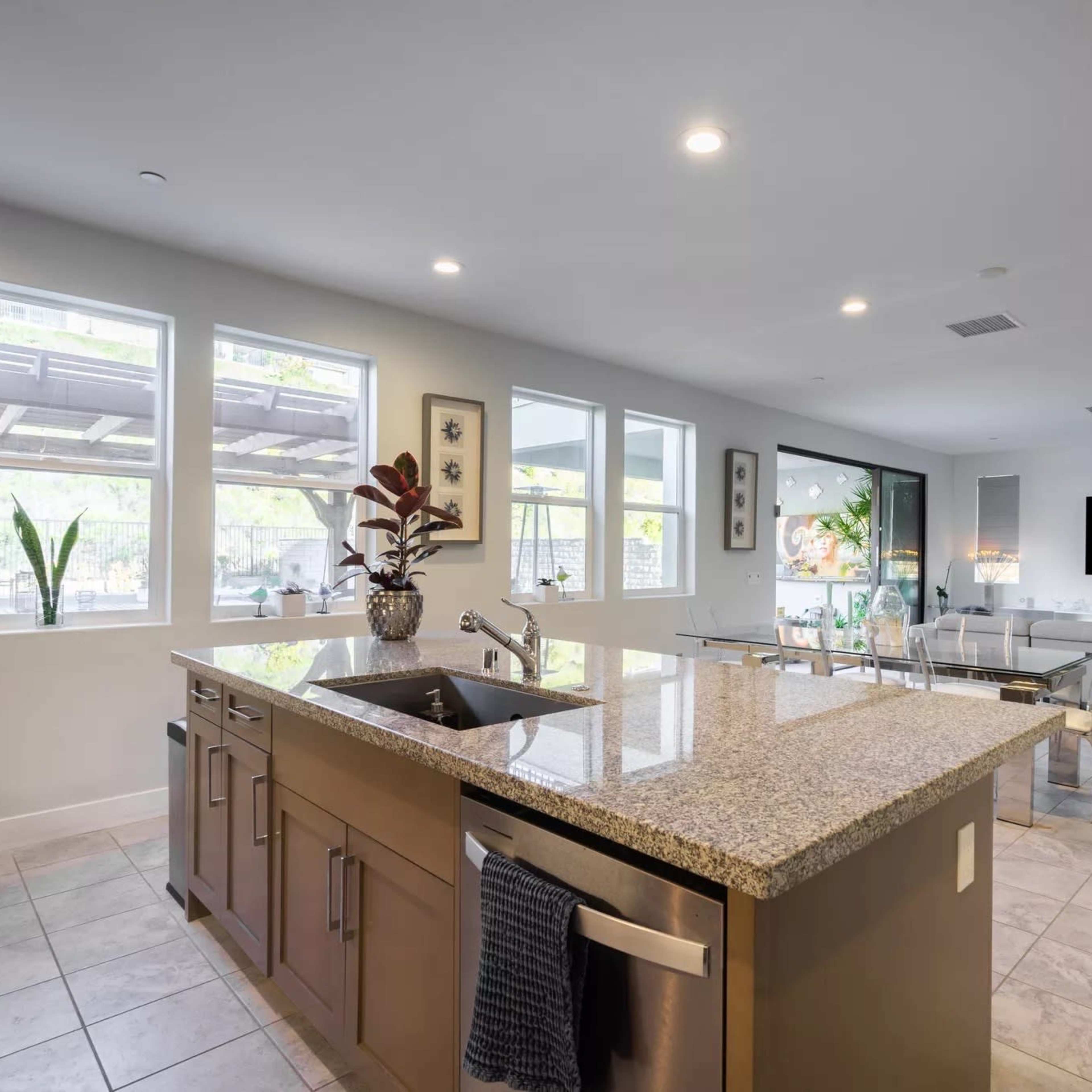 A modern kitchen with a granite countertop, featuring a sink and appliances, adjacent to a dining area with large windows and natural light.