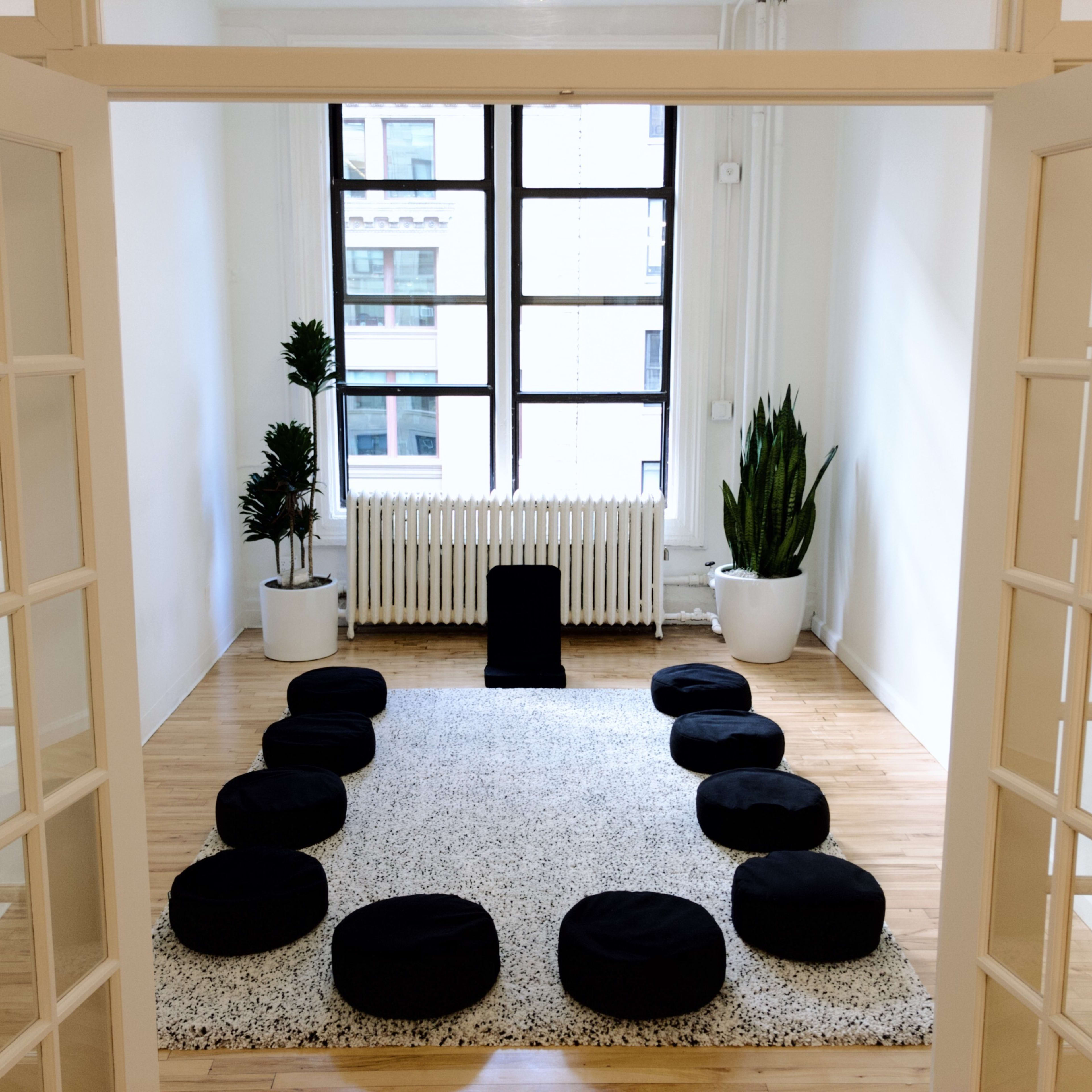 A spacious, bright room with a circular arrangement of black cushions on a light-colored rug, flanked by plants and a radiator below large windows.