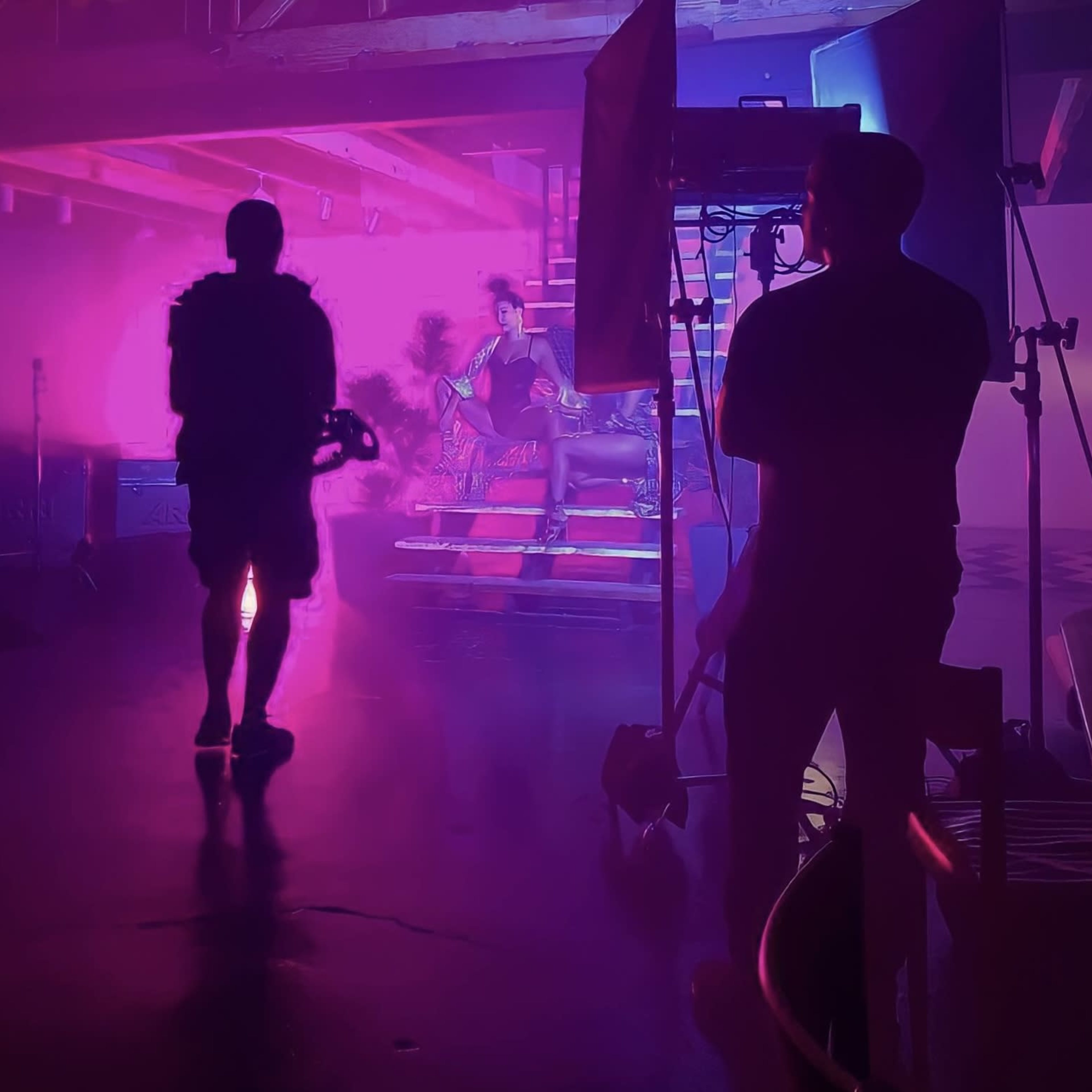 A photoshoot is taking place in a dimly lit studio with colorful lighting, where a subject is posed on a staircase while crew members adjust equipment.