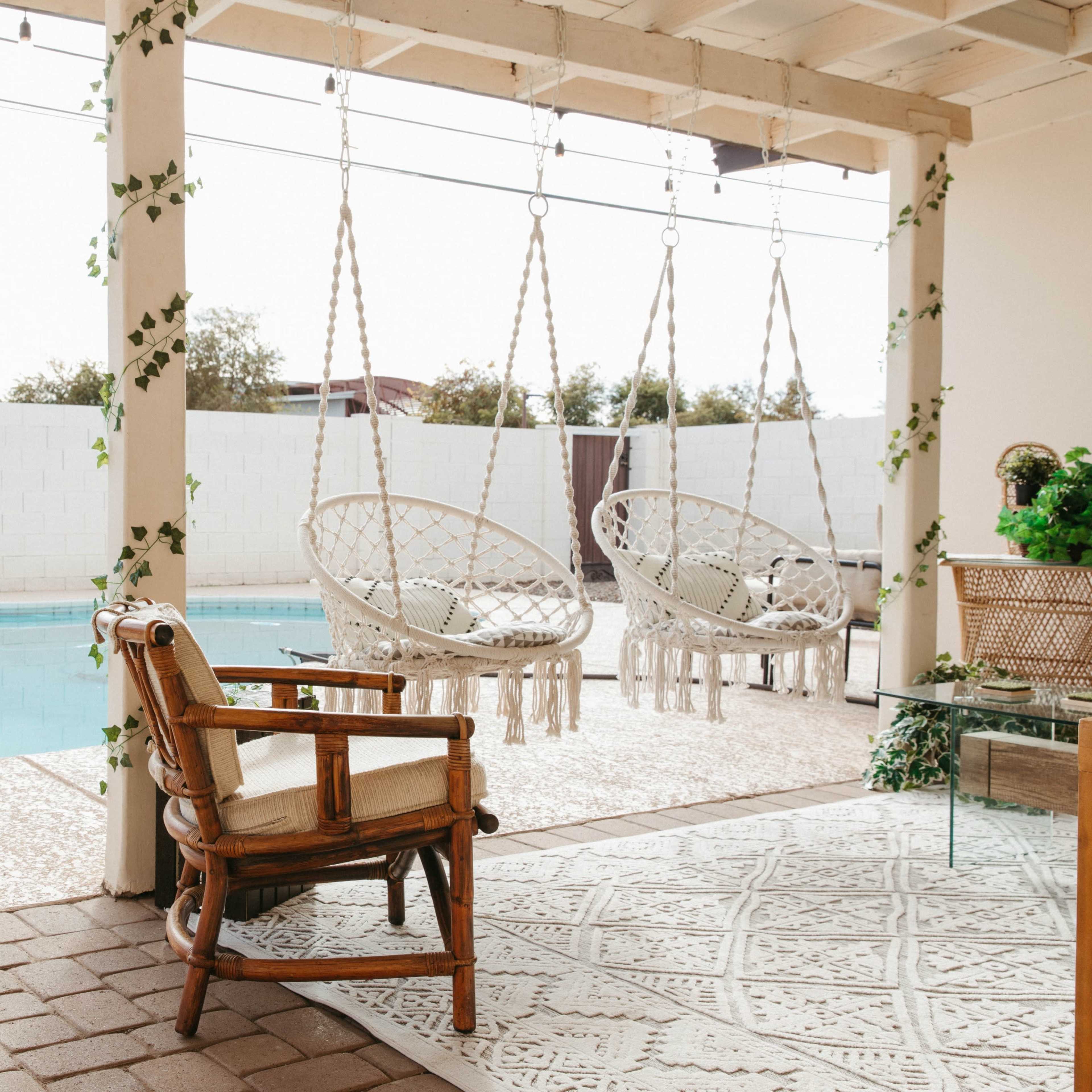 A pair of hanging wicker chairs are positioned next to a swimming pool, with a wooden chair and patterned rug nearby.