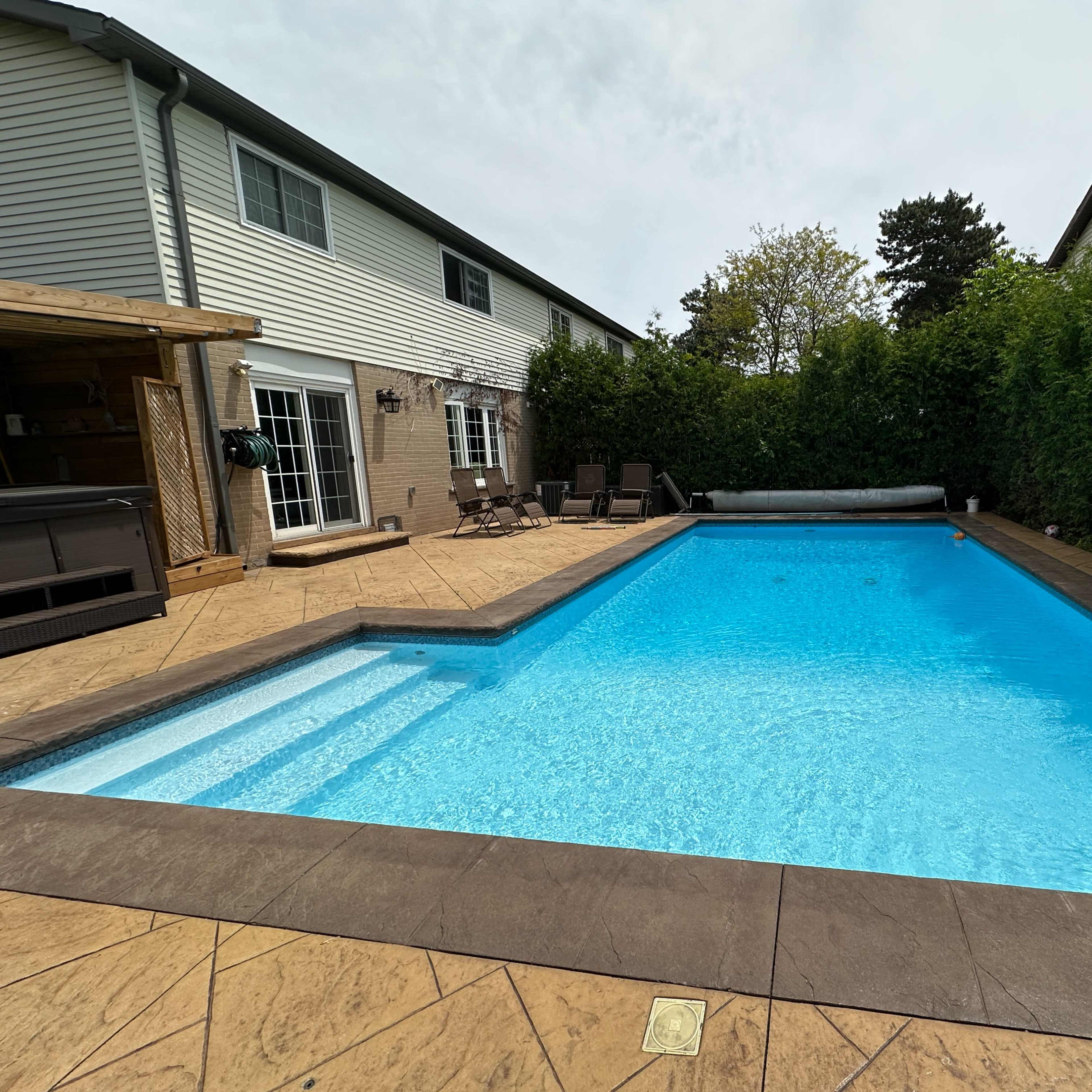 A rectangular swimming pool surrounded by a stone deck and bordered by tall hedges, with a house visible in the background.