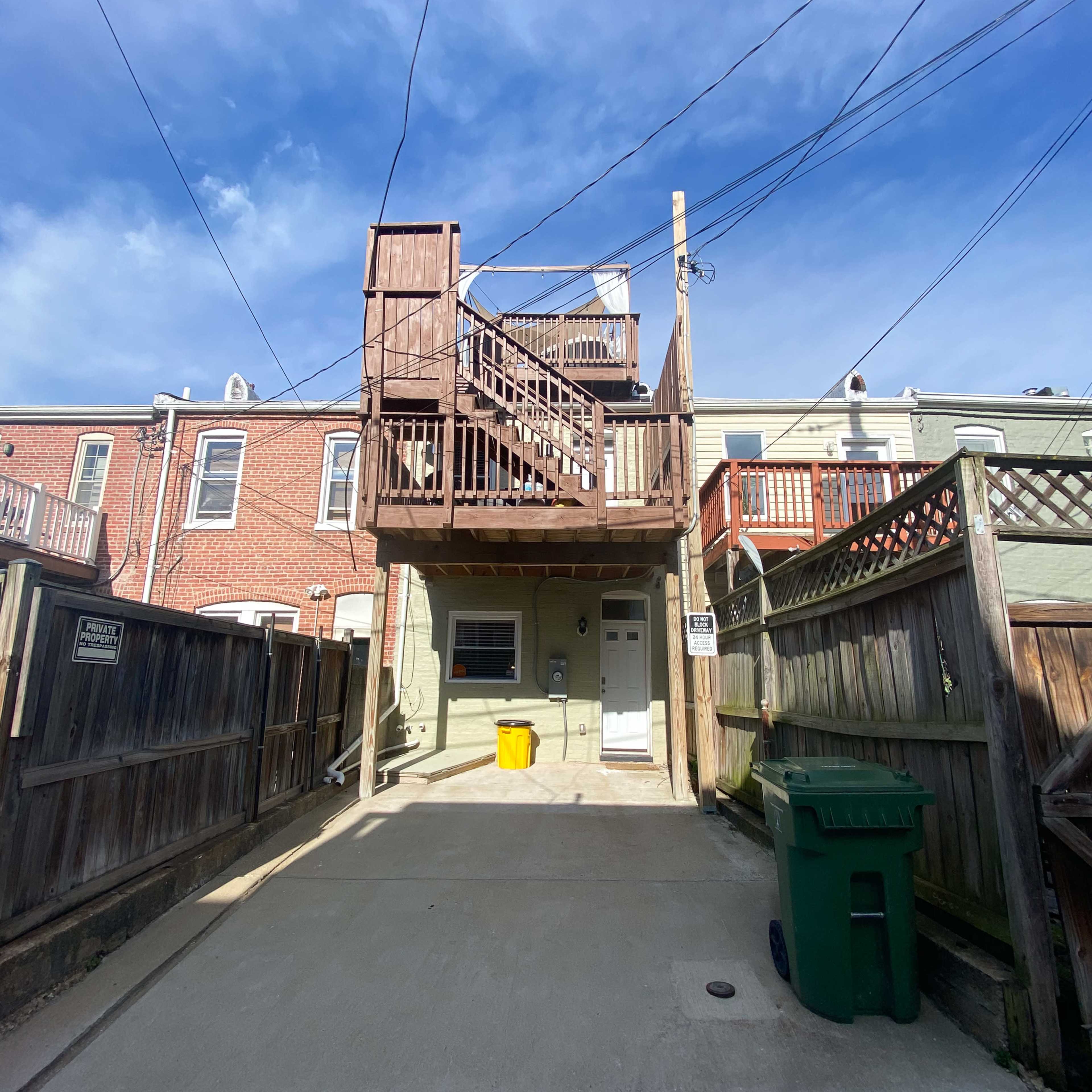 A narrow alleyway with a two-story wooden structure at the back, lined with power lines, and a green trash can on the right.
